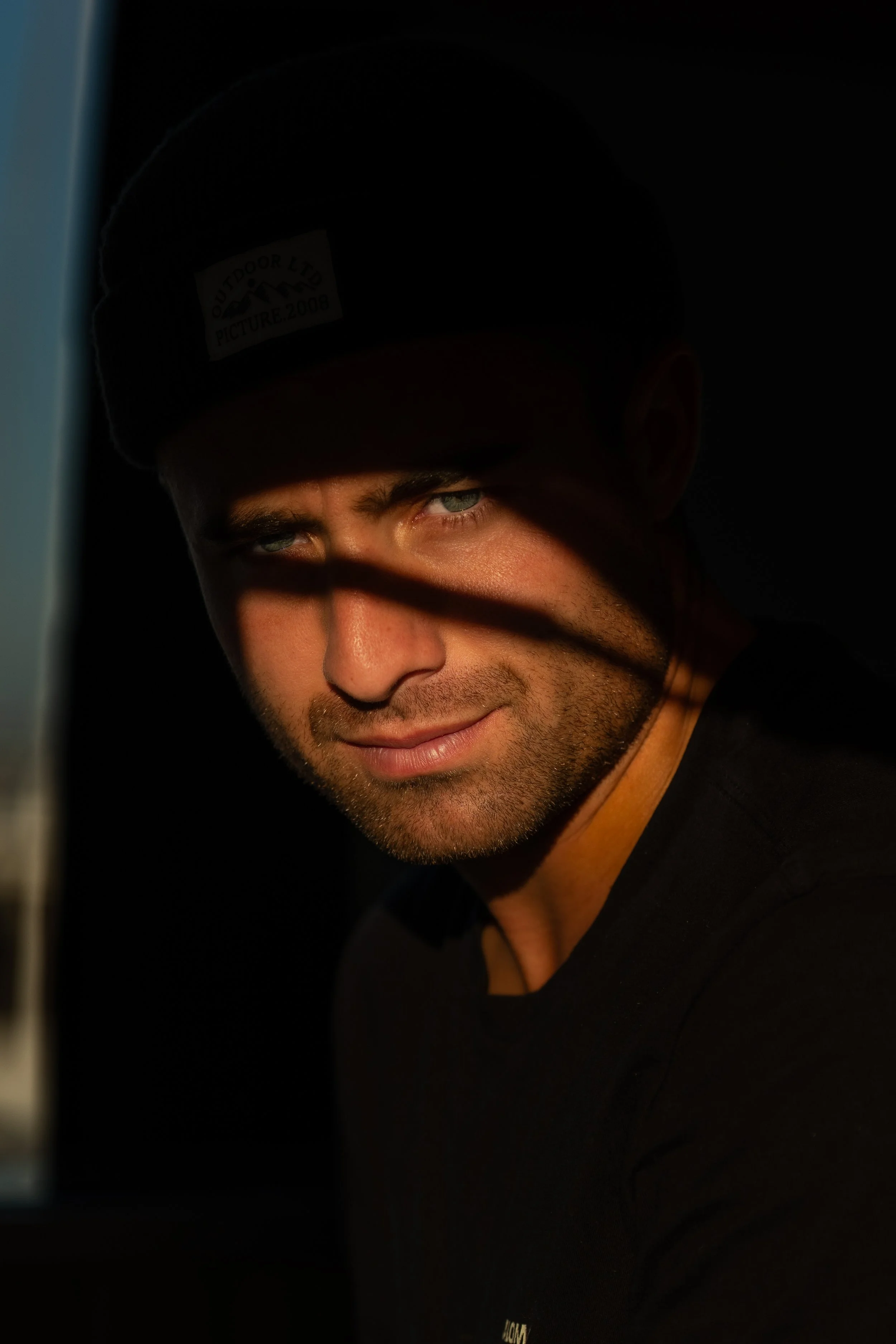 Un homme avec des yeux bleus regarde vers la caméra, partiellement dans l'ombre avec une lumière chaude qui illumine son visage.