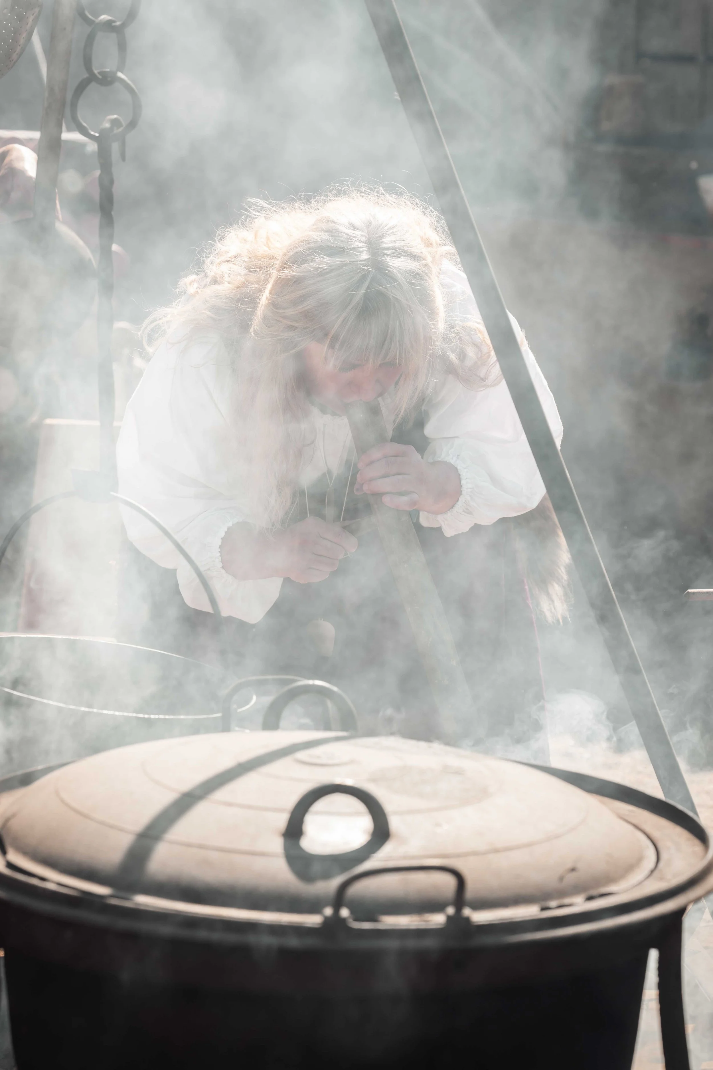 Une femme déguisée en sorcière ou en personnage de conte de fées, avec des cheveux blonds et longs, regarde dans un chaudron en train de fumer ou de cuire quelque chose.