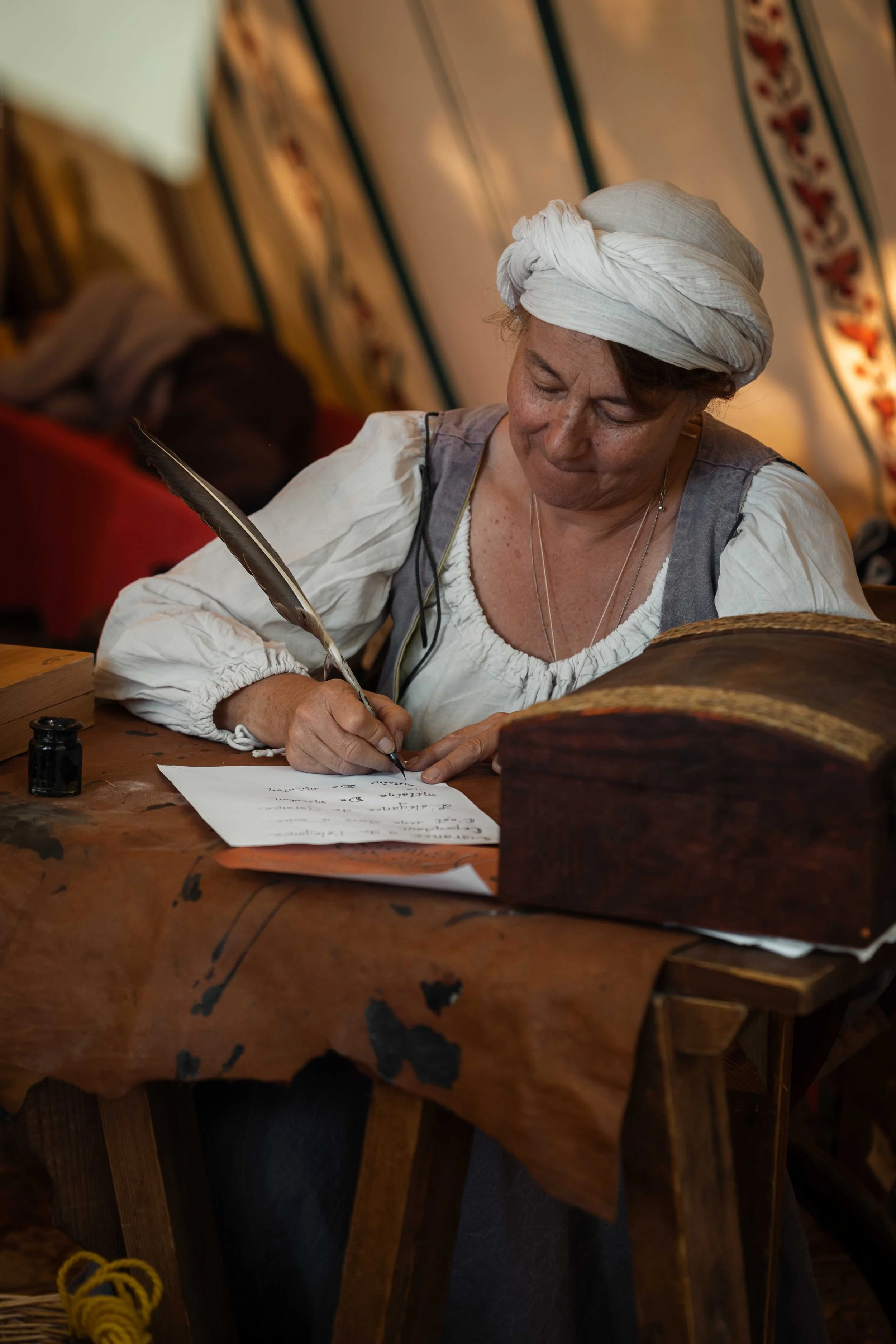Femme habillée en costume traditionnel médiéval, portant un turban blanc, écrivant à la plume sur une feuille, dans une ambiance ancienne avec une table en bois et un coffre en bois.