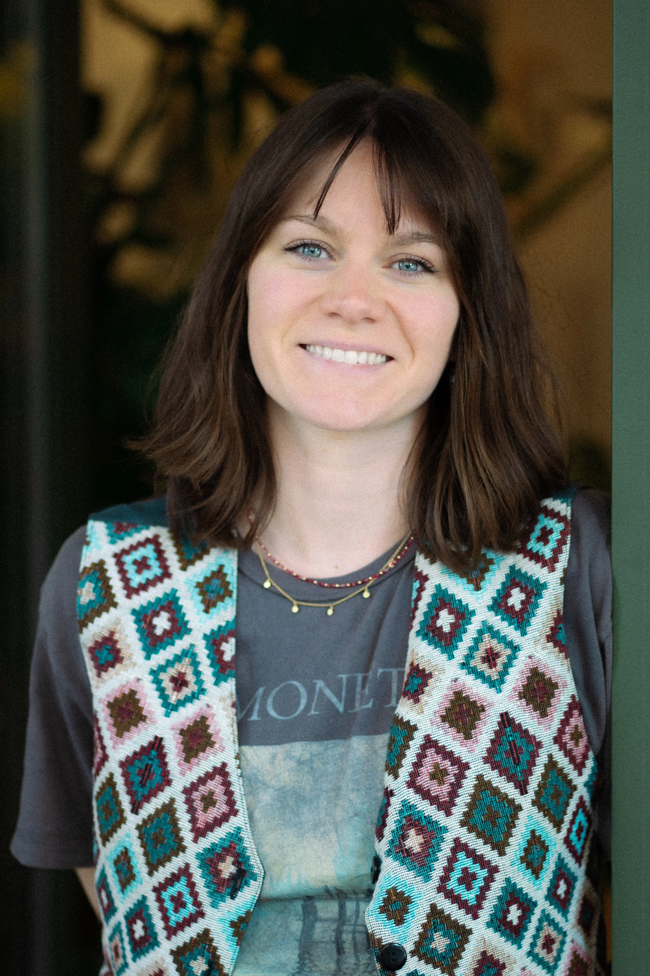 Jeune femme avec des cheveux châtains, habillée d'un t-shirt gris et d'une veste avec un motif géométrique coloré, souriant dans un environnement intérieur.