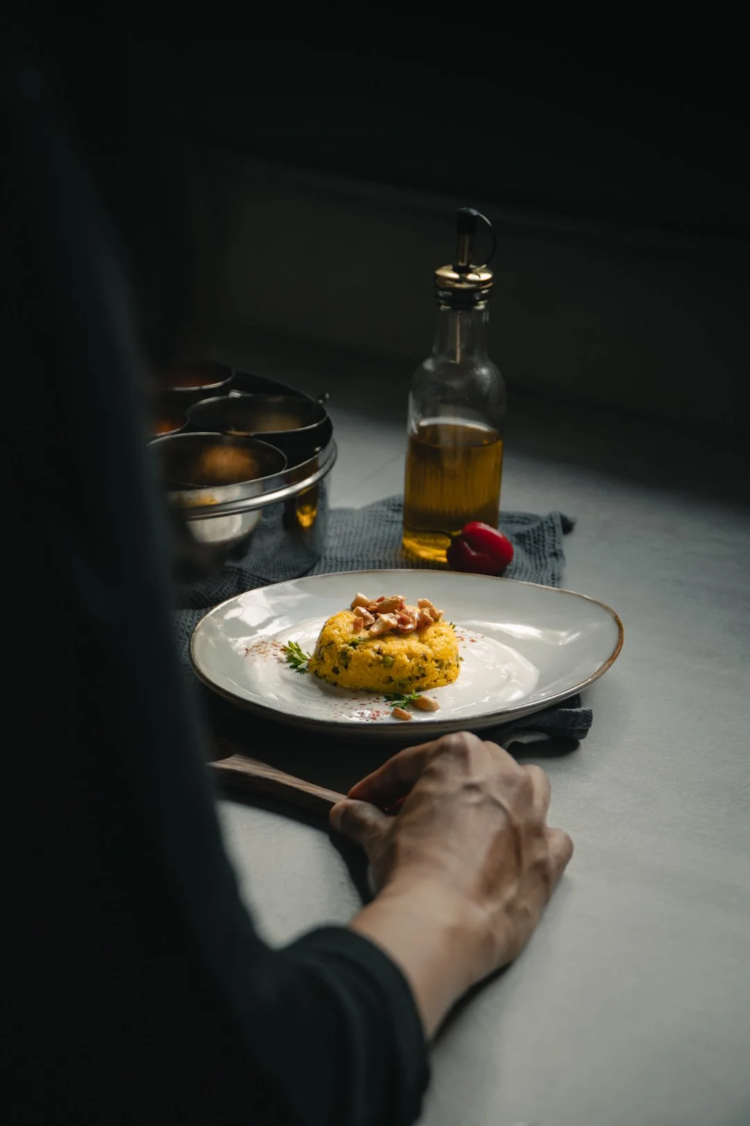 Assiette de polenta servie avec de l'huile d'olive et des noix, accompagnée d'un poivron rouge et d'herbes, sur une nappe noire. En arrière-plan, un flacon d'huile d'olive, un bol noir, un bol blanc et un couteau posé sur la table.