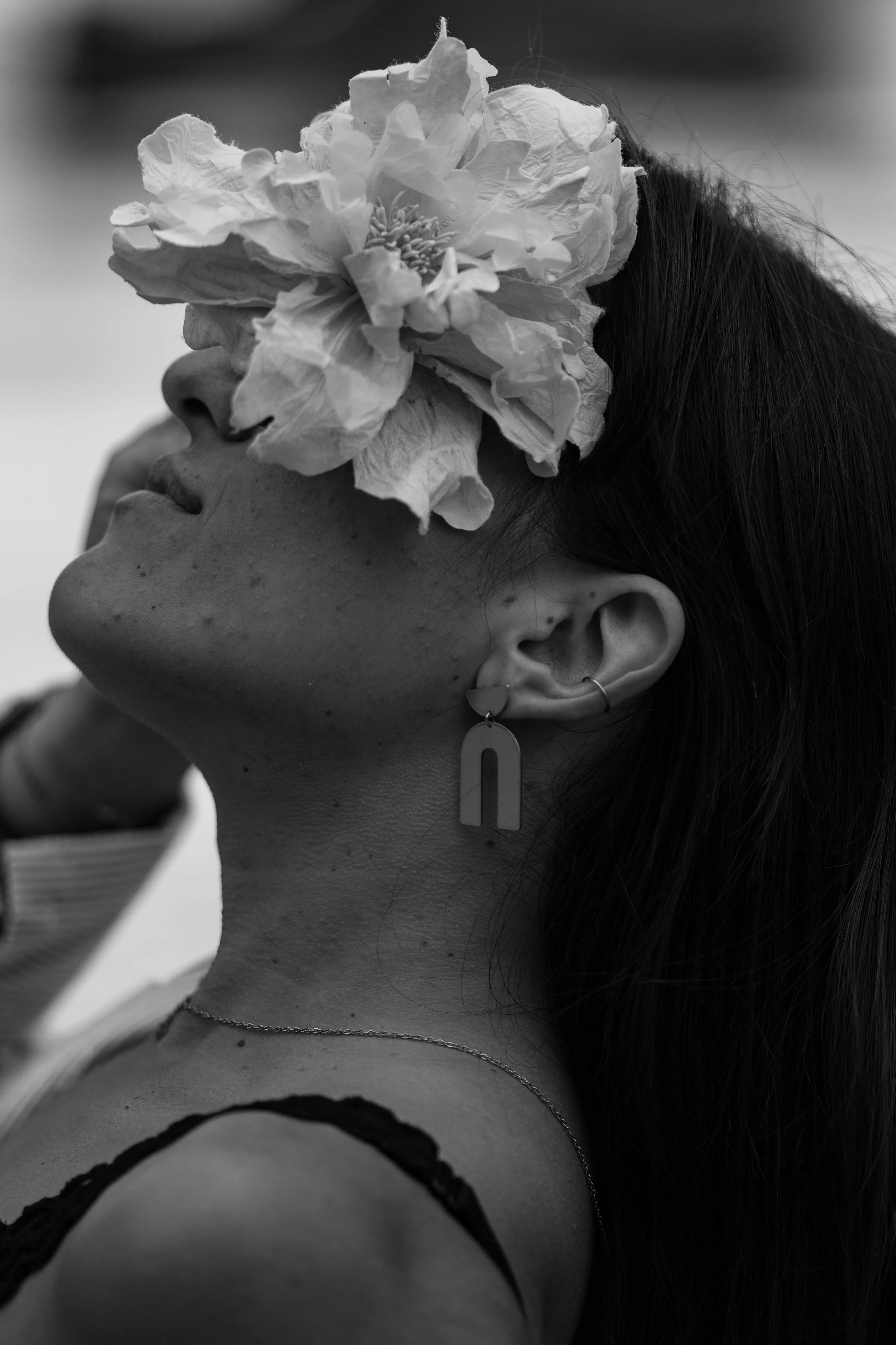 Portrait d'une femme avec un grand fleur sur l’œil, portée en noir et blanc, portant des boucles d'oreilles modernes et un collier, coiffée de cheveux longs.