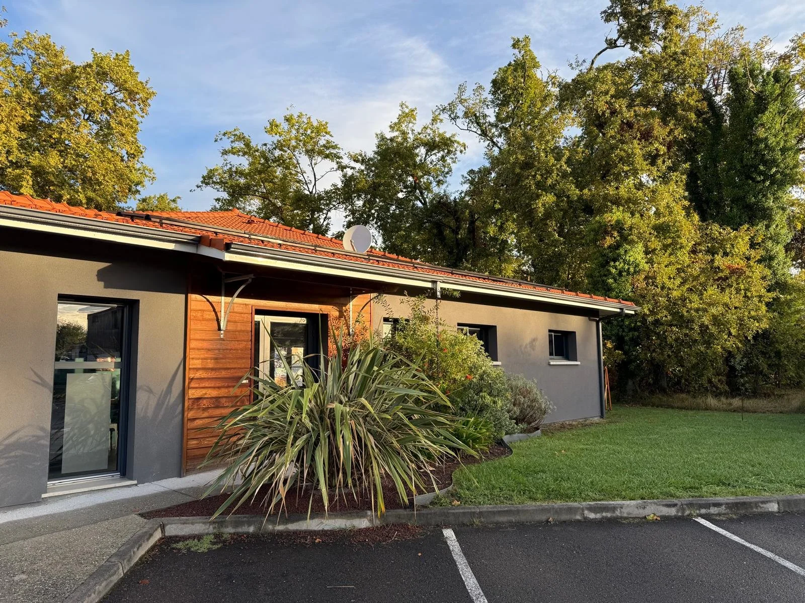 Une maison moderne avec un mur gris, un toit en tuiles rouges, entourée d'arbres et de végétation, avec une zone de stationnement vide en avant.