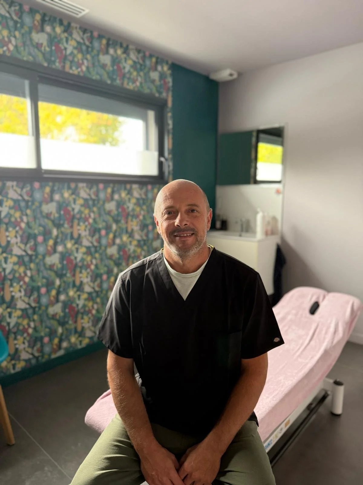 Un professionnel de la santé souriant, assis sur une table de massage dans une salle lumineuse avec une décoration murale colorée.