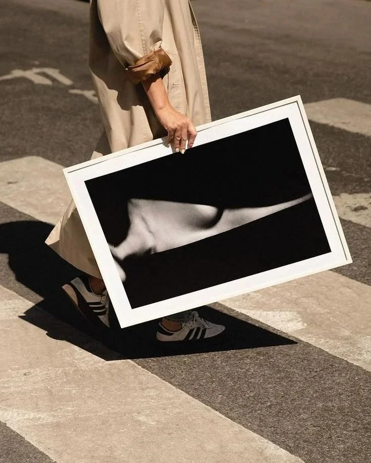 Person, die ein schwarzes und weißes Kunstwerk auf dem Boden hält, das eine menschliche Körperlinie zeigt, während sie auf einer Straße mit Zebrastreifen geht.