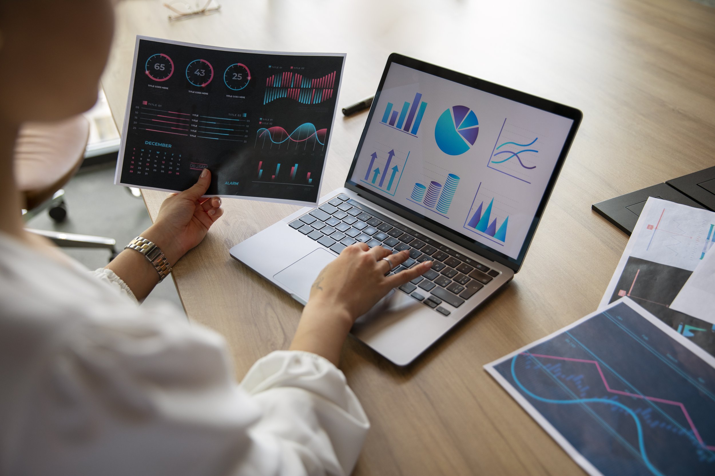 Person working at a desk with a laptop displaying various colorful graphs and charts, holding a printed sheet with similar data visualizations.
