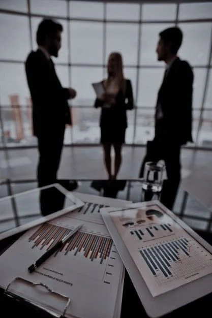 Business meeting scene with three professionals standing behind a conference table, discussing in front of large windows. The table has documents, charts, a tablet, and a glass of water.