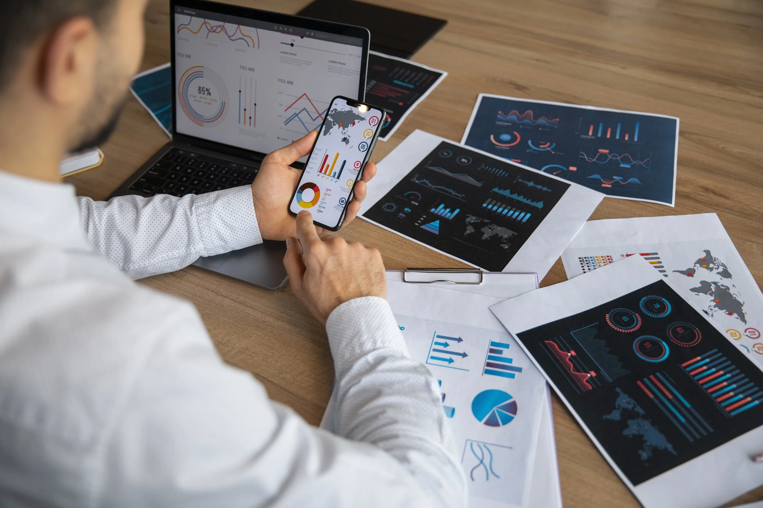 A person working on data analysis, using a smartphone and laptop surrounded by printed charts and graphs with various data visualizations on a wooden desk.