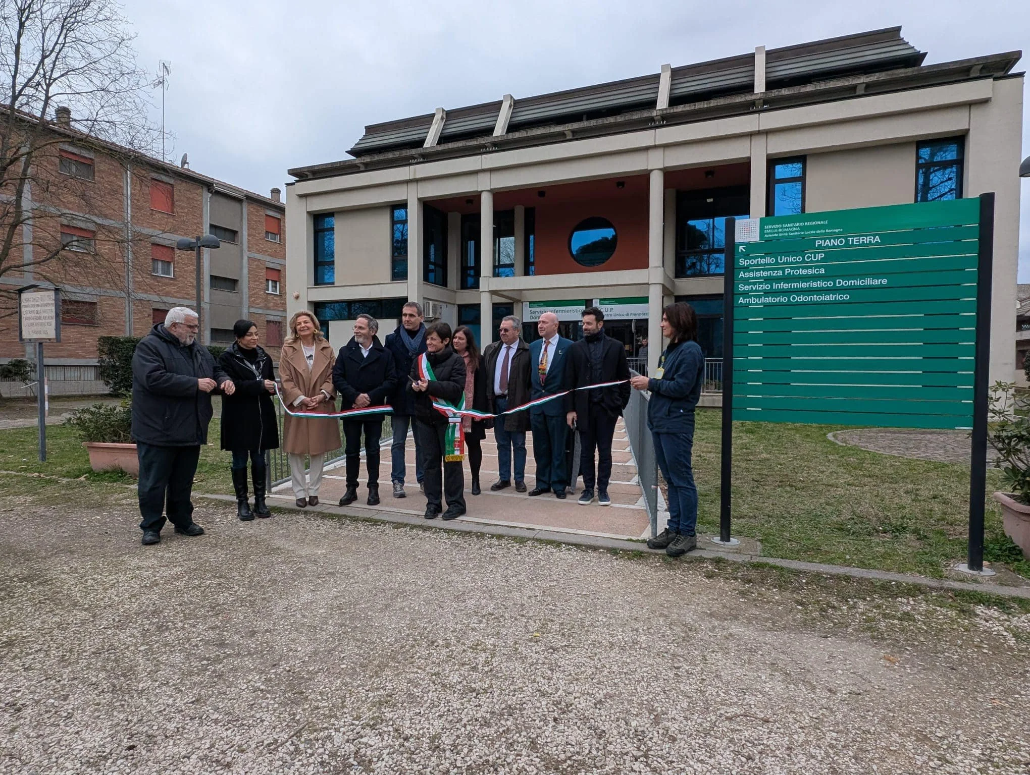 Gruppo di persone durante una cerimonia di inaugurazione davanti a un edificio pubblico, con un nastro da tagliare e un cartello che indica vari servizi sanitari
