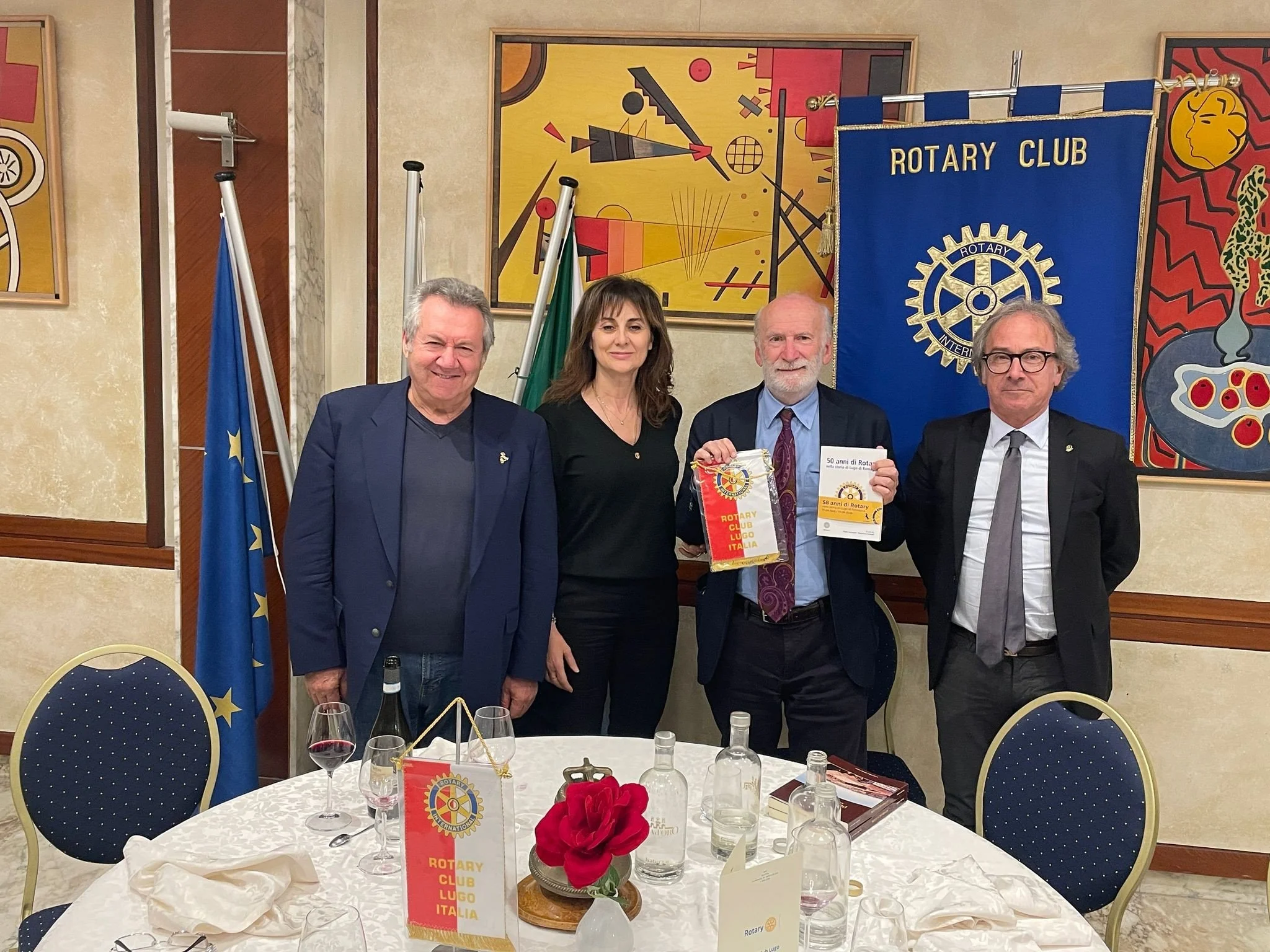 Quattro persone posano per una foto in una sala con quadri astratti alle pareti e una bandiera blu del Rotary Club. Due uomini sullo sfondo, una donna al centro e un uomo che tiene un libro e un sacchetto con il logo del Rotary, tutte con abiti forma