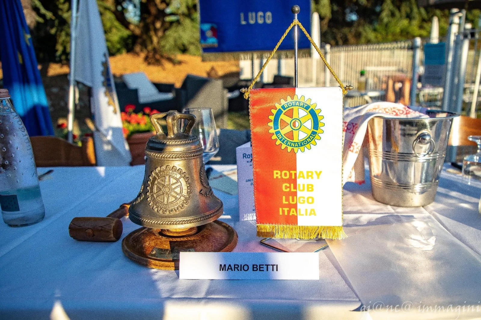 Tavola con distintivo Rotary Club Lugo Italia, campana in metallo, bicchieri d'acqua, bottiglia d'acqua, secchiello in metallo, tovaglioli, e un nome 'Mario Betti' sulla tavola.
