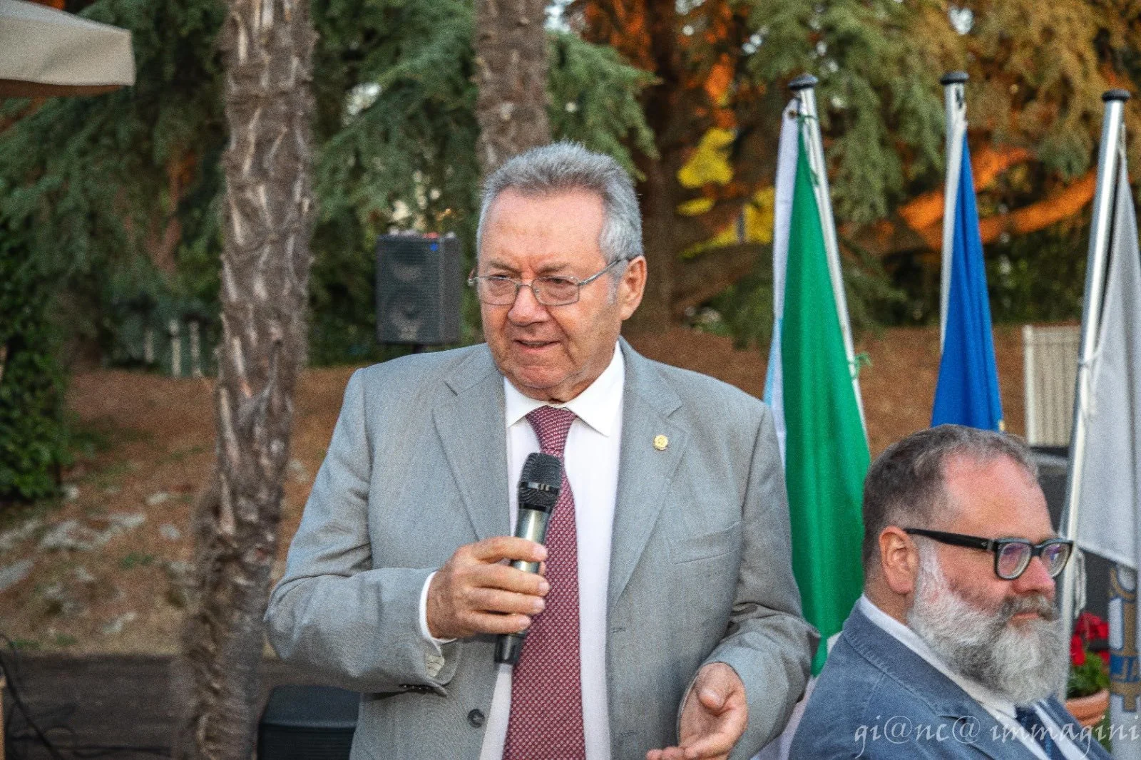 Un uomo anziano con occhiali, giacca grigia e cravatta rosa che tiene un microfono mentre parla, con bandiere italiane e europee sullo sfondo, durante un evento all'aperto.