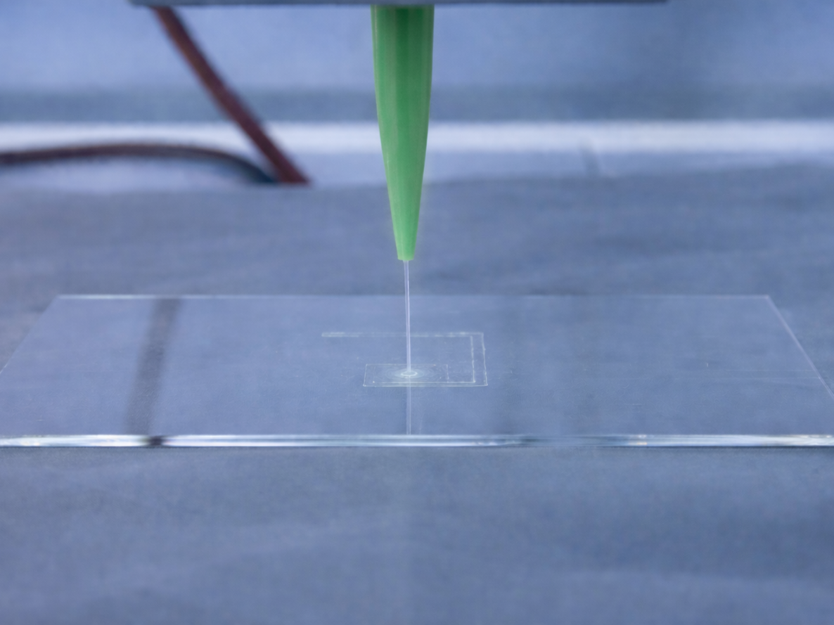 Close-up of a pipette dispensing a liquid droplet onto a small, transparent, square glass slide on a metallic surface.