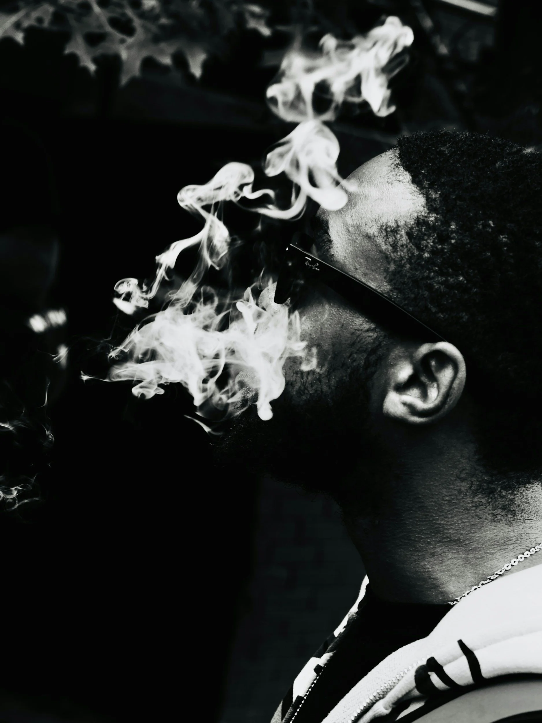 A person with dark, curly hair and sunglasses exhaling smoke, wearing a white jacket with black accents and a chain necklace.
