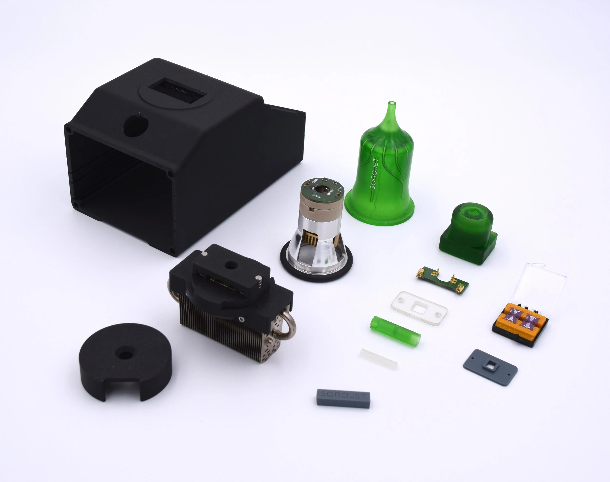 Collection of electronic and mechanical components on a white background, including a black rectangular device, a green plastic cone, a small metallic component, and various small circuit parts.