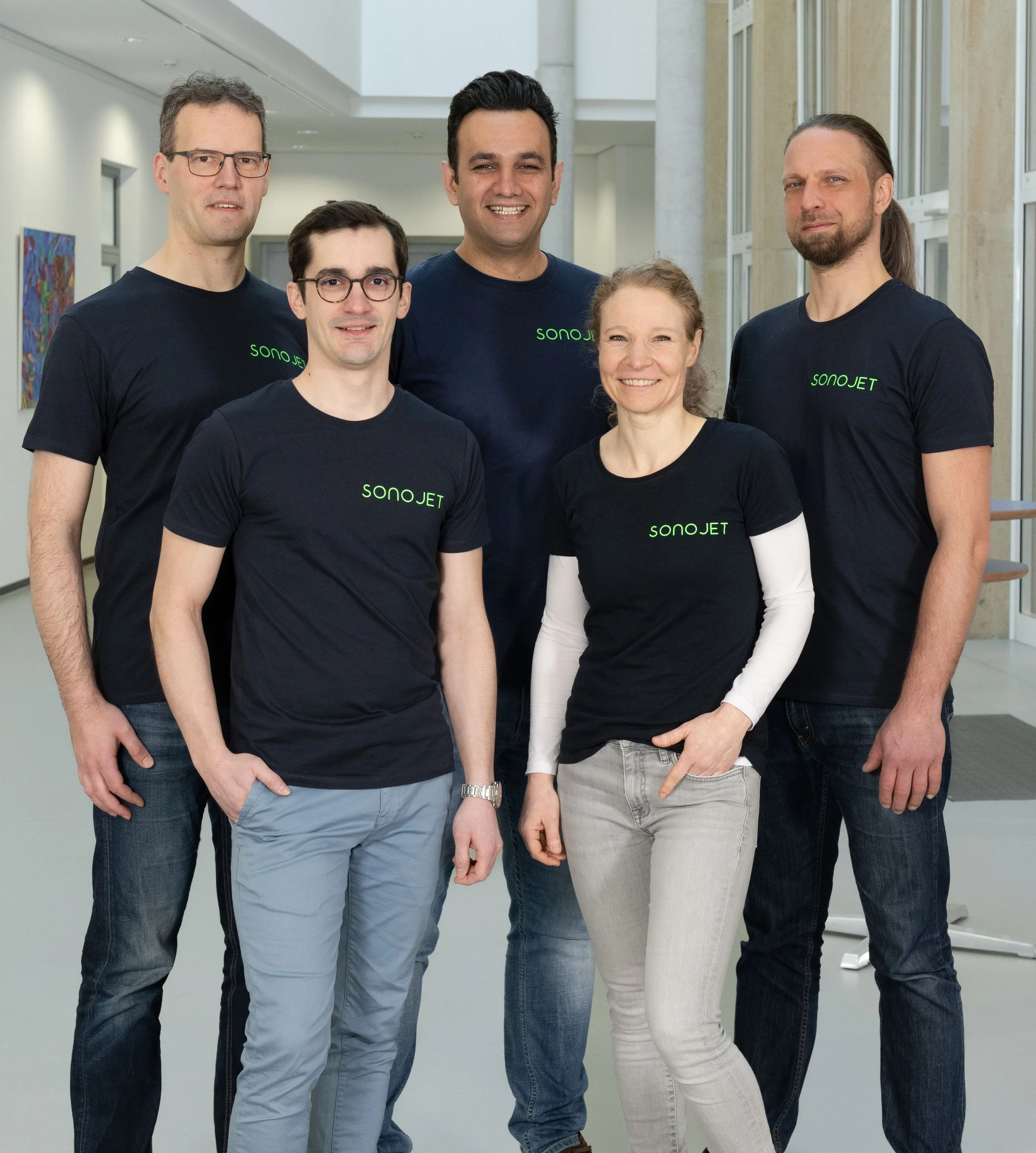 A diverse group of five people, three men and two women, standing in an indoor setting with large windows and artwork on the wall, all wearing navy blue T-shirts with the word 'SONOJET' printed on them, smiling and posing for the photo.