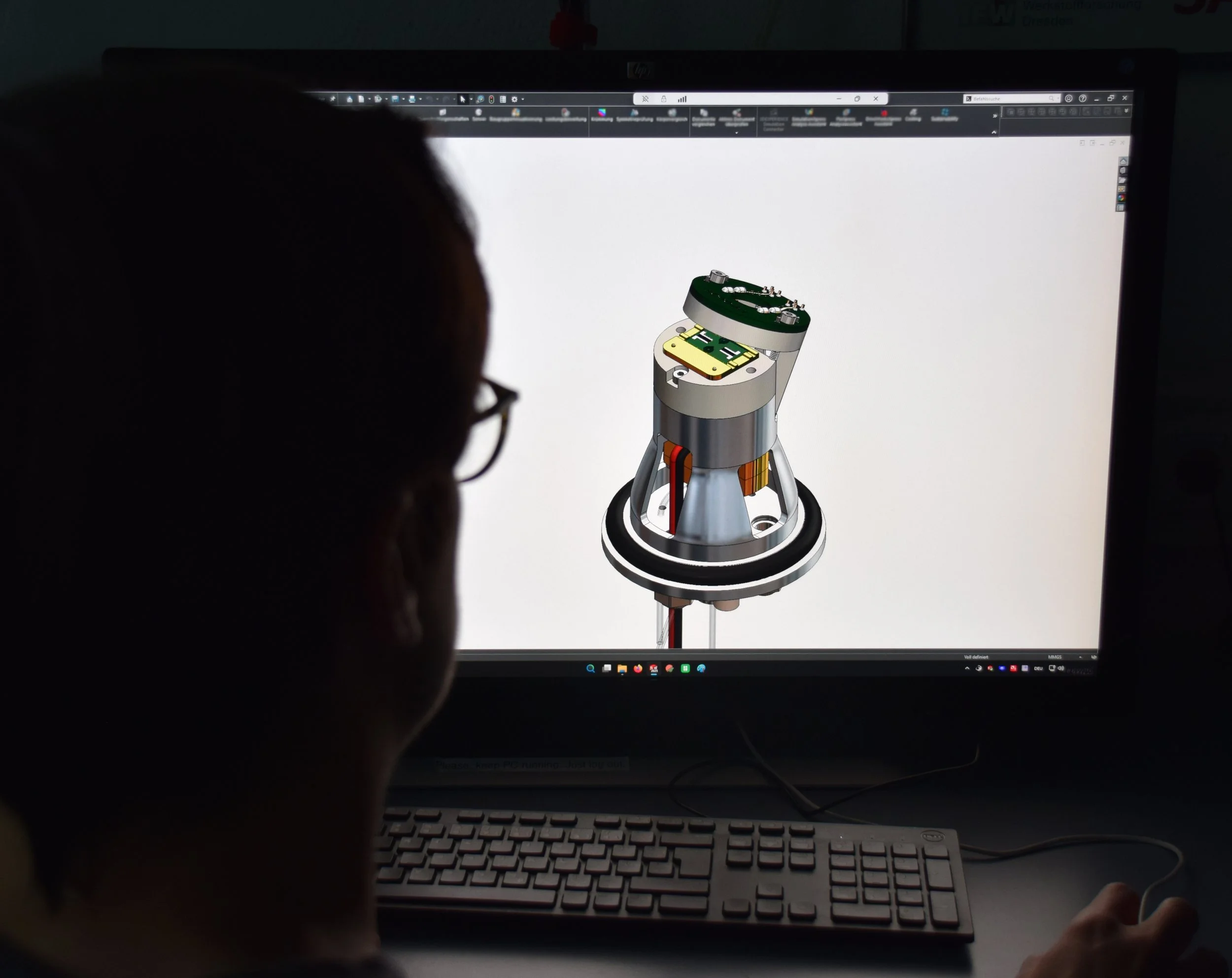 A computer screen displaying a 3D model of a mechanical component or device, with a person's silhouette visible in the foreground.