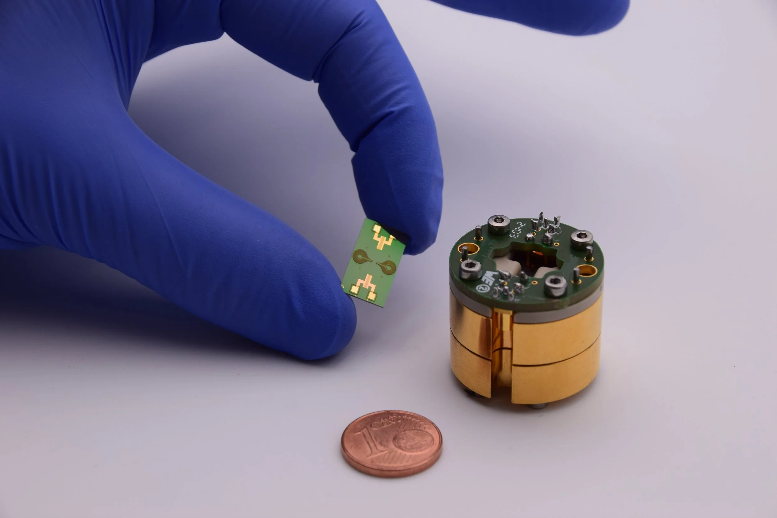 A person wearing blue gloves handling a small green electronic circuit with gold contacts, above a copper-colored cylindrical component, with a penny coin nearby for size reference.