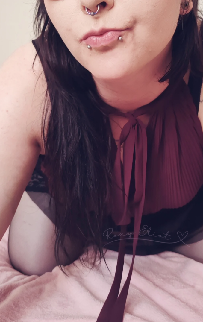 Close-up of a femme person with dark hair, multiple facial piercings, wearing a burgundy top with a bow, leaning forward on a pink blanket.