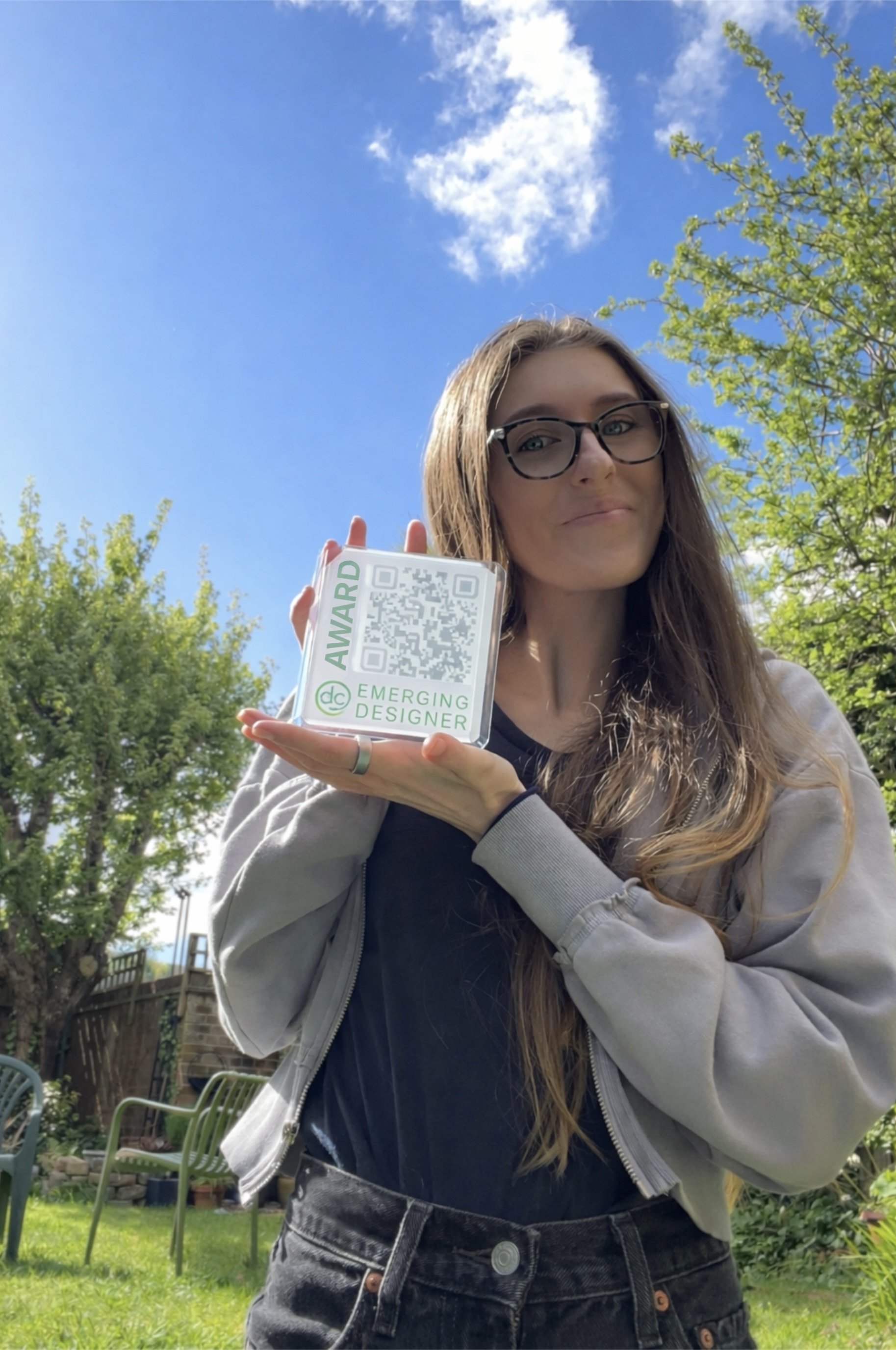 A young woman with long hair, glasses, and a smile standing outdoors on a sunny day, holding a badge that reads 'AWARD' and 'Emerging Designer' with a QR code. She is wearing a jacket over a dark shirt, with green trees and blue sky in the background.