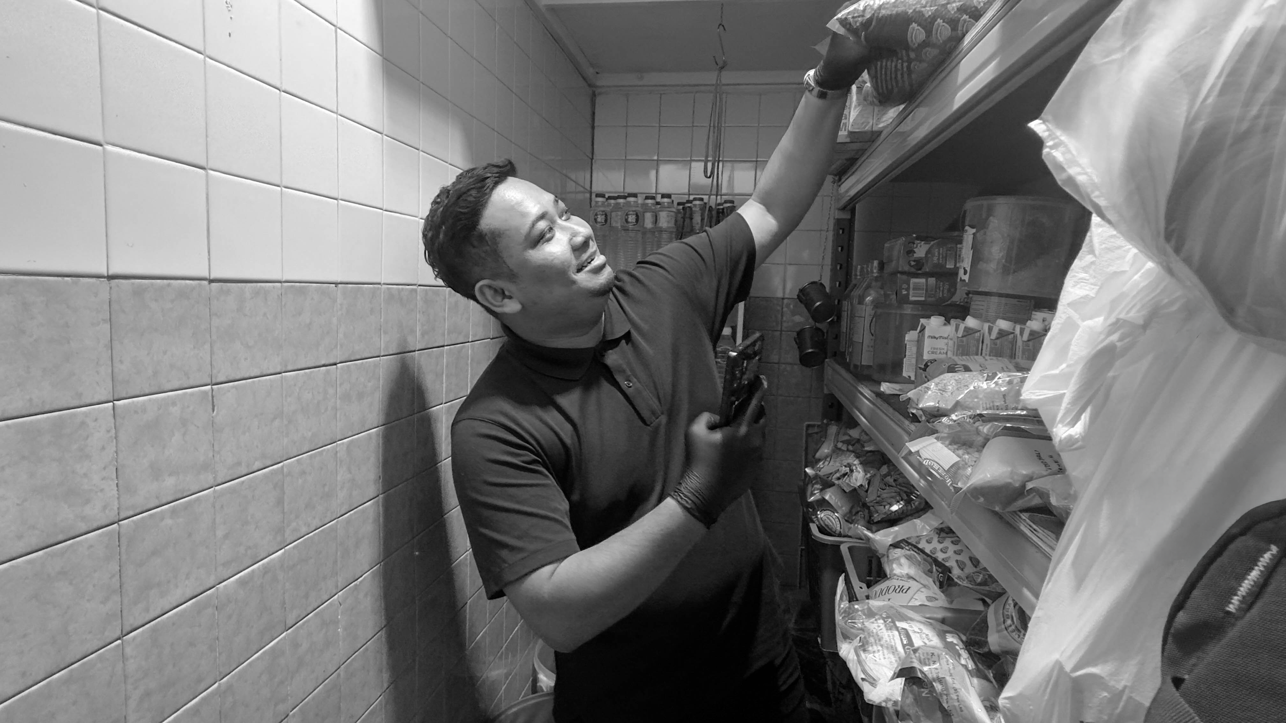 A man wearing a polo shirt and gloves shopping in a grocery store, reaching for an item on a shelf.