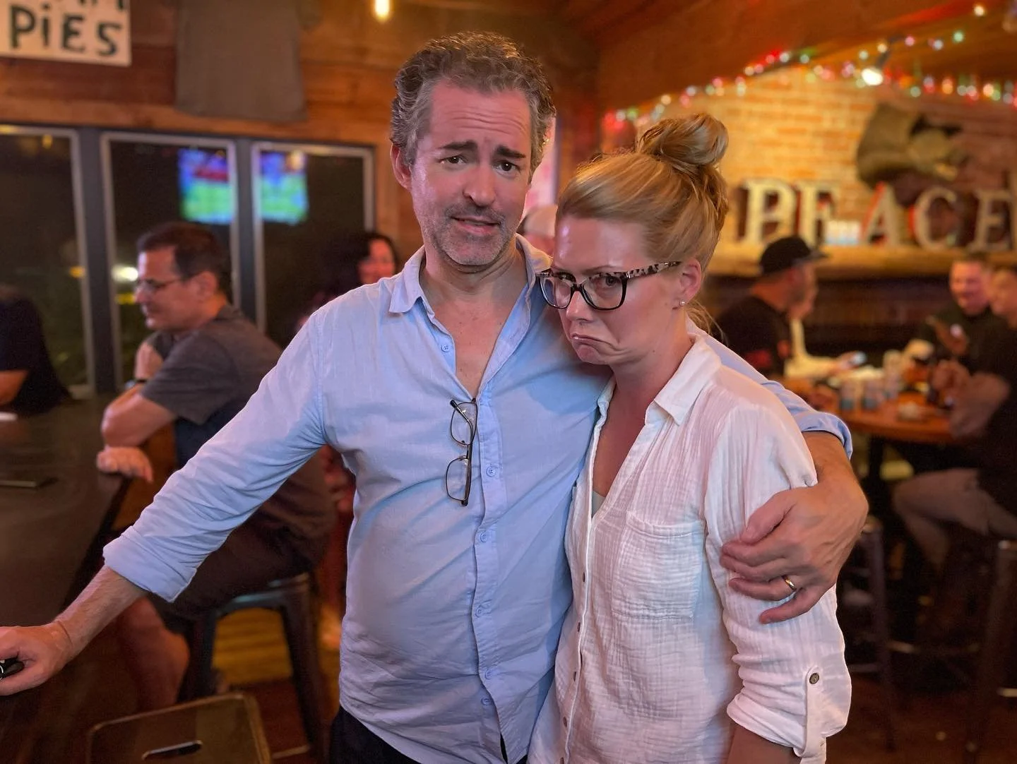 A man and woman with humorous facial expressions in a bar. The man has gray, curly hair, a beard, and is wearing a light blue shirt with glasses hanging from it. The woman is wearing glasses, has her hair in a bun, and is dressed in a white shirt. Th