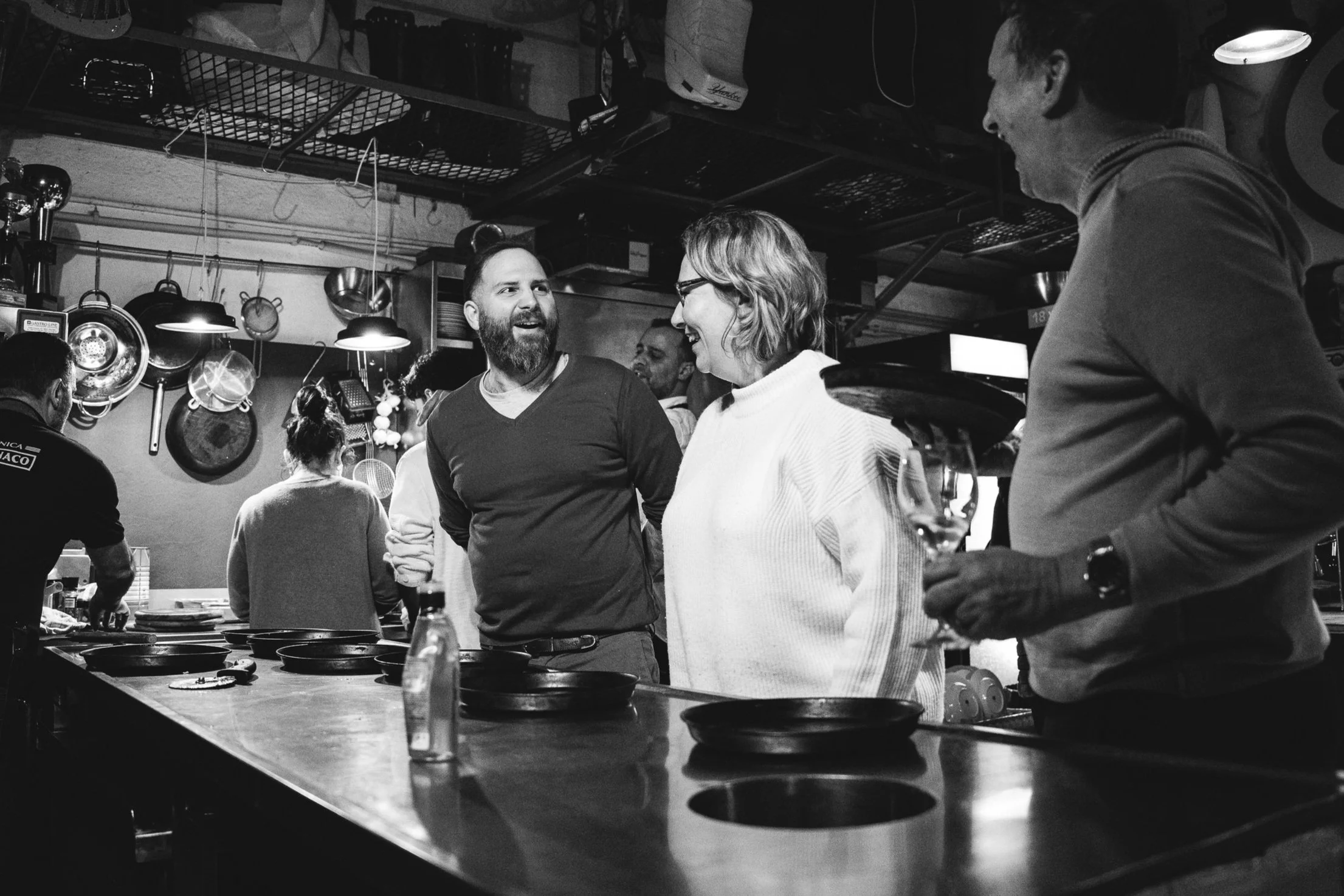 Menschen unterhalten sich in einer Bar oder einem Restaurant, während neben ihnen ein Kellner Getränke serviert. Die Szene ist schwarz-weiß und zeigt eine lebhafte Atmosphäre.