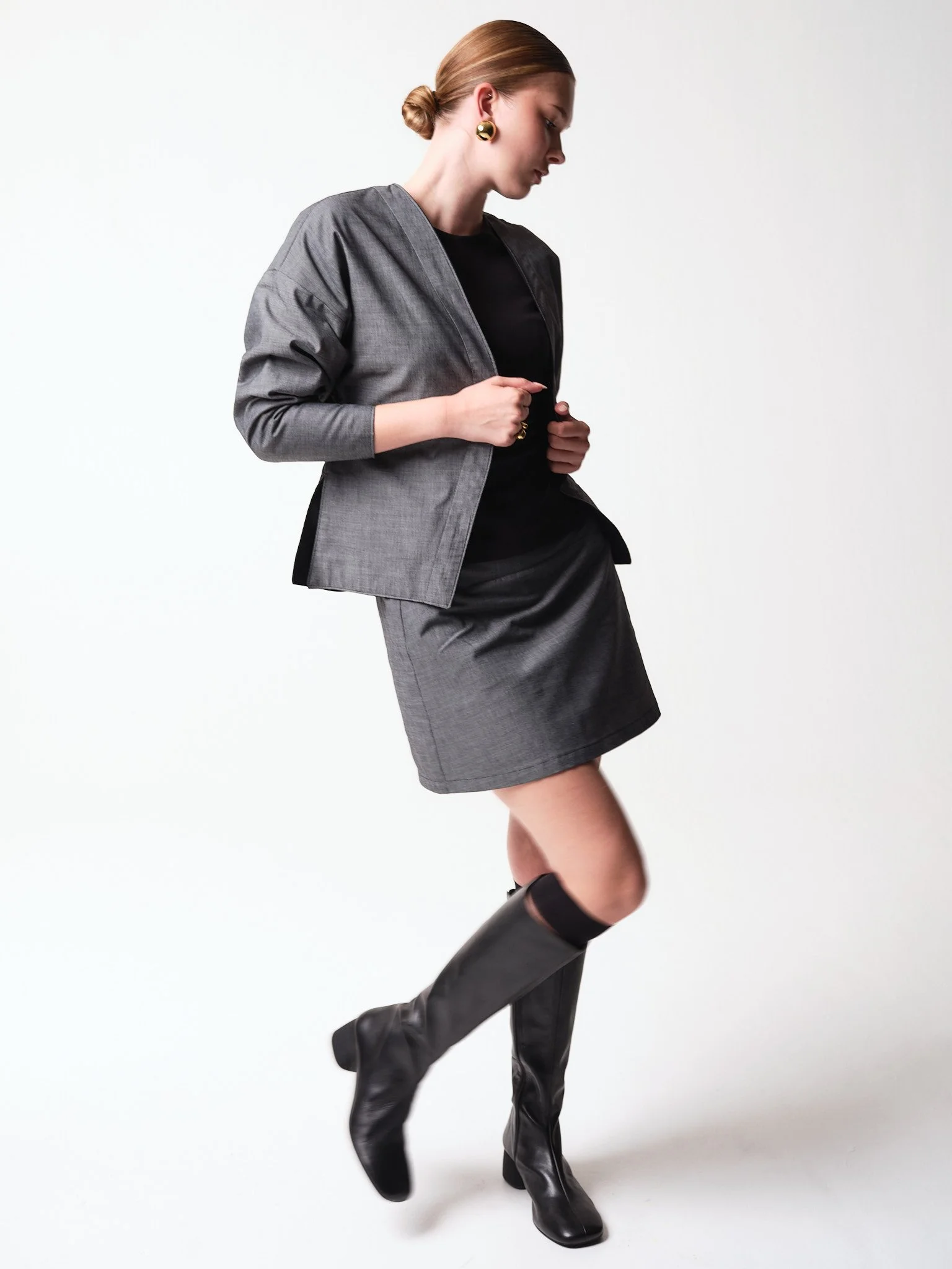 Frau trägt graue Jacke, schwarzen Oberteil, grauen Rock und schwarze Stiefel auf weißem Hintergrund.