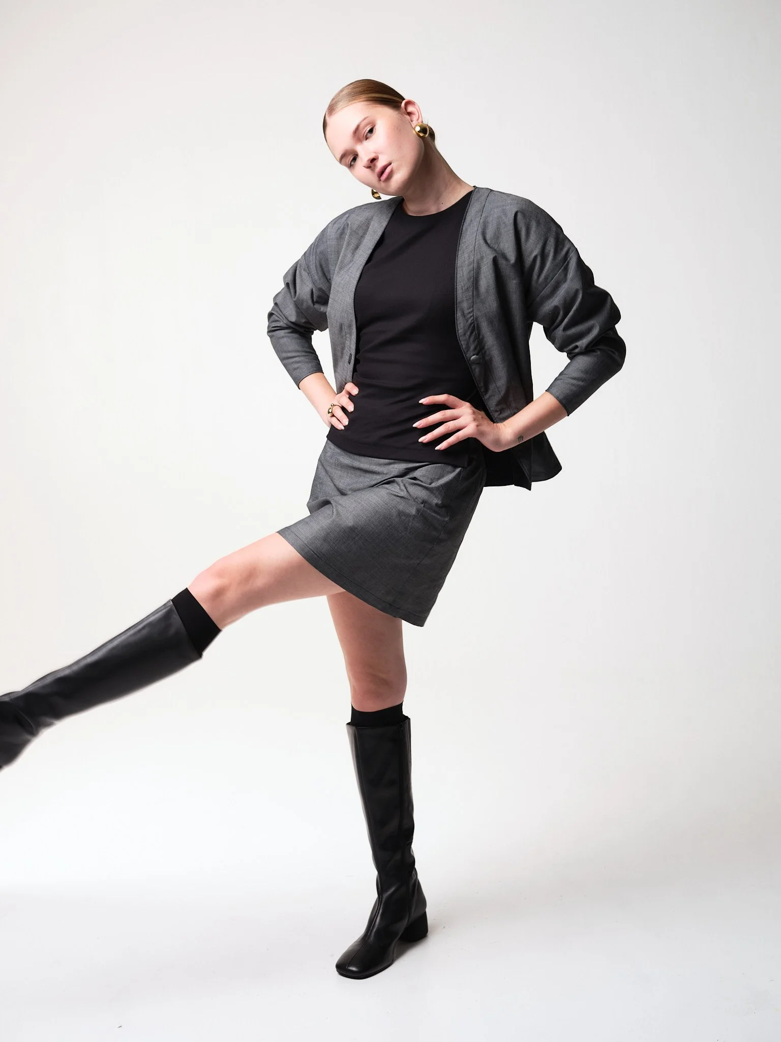 Eine modebewusste Frau trägt ein schwarzes Top, einen grauen Blazer und einen grauen Rock, kombiniert mit schwarzen hohen Stiefeln, und posiert vor einem weißen Hintergrund.