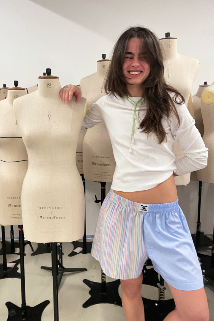 Ung kvinde med langt brunt hår, smilende, står i en kreativ tøjdesign-klasseværelse blandt mannequins, iført en hvid trøje og farverige bæredygtige shorts i København Danmark
