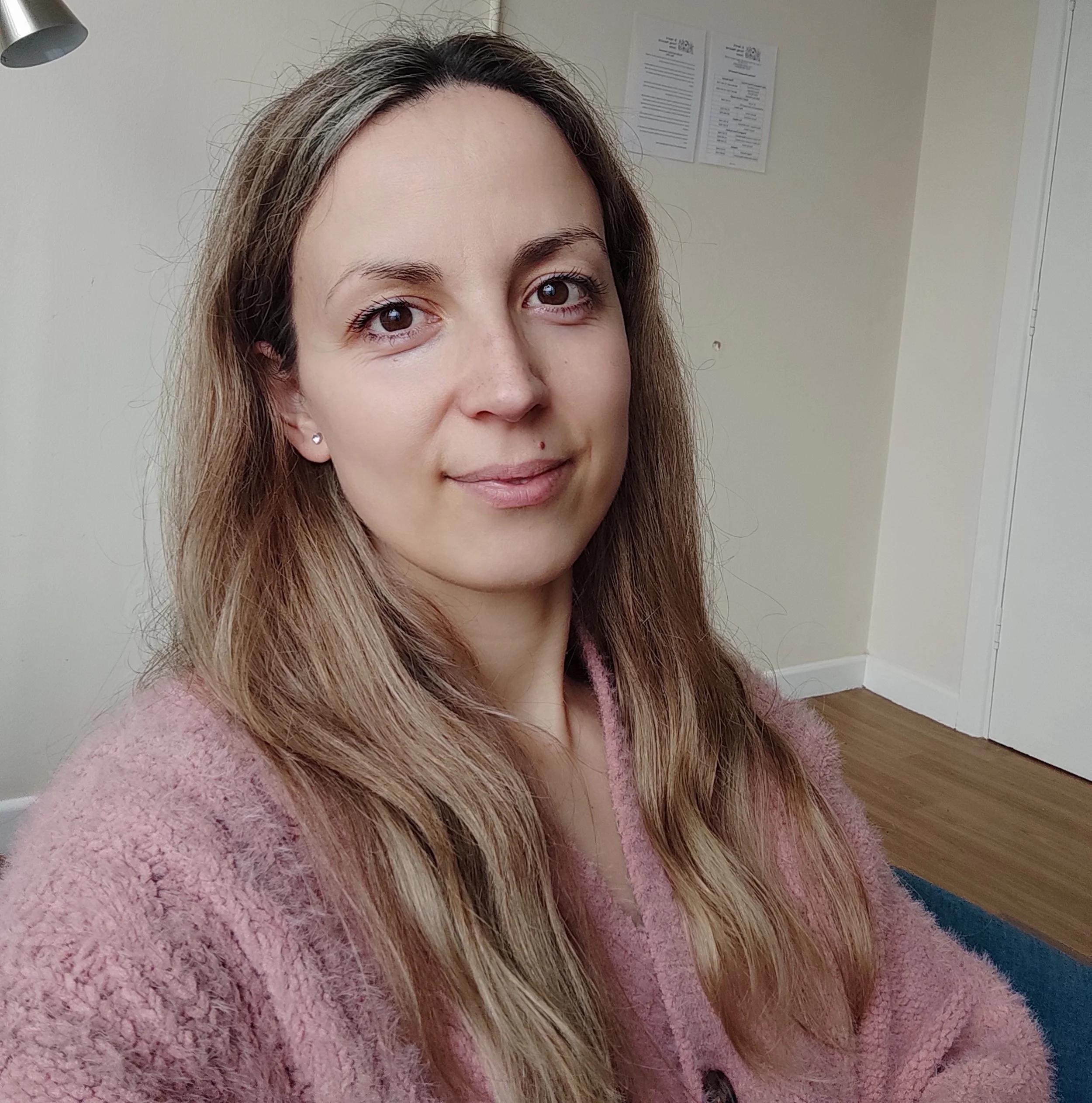 A woman with long light brown hair and light skin looking at the camera, wearing a pink fuzzy sweater, in a room with beige walls and wooden flooring.