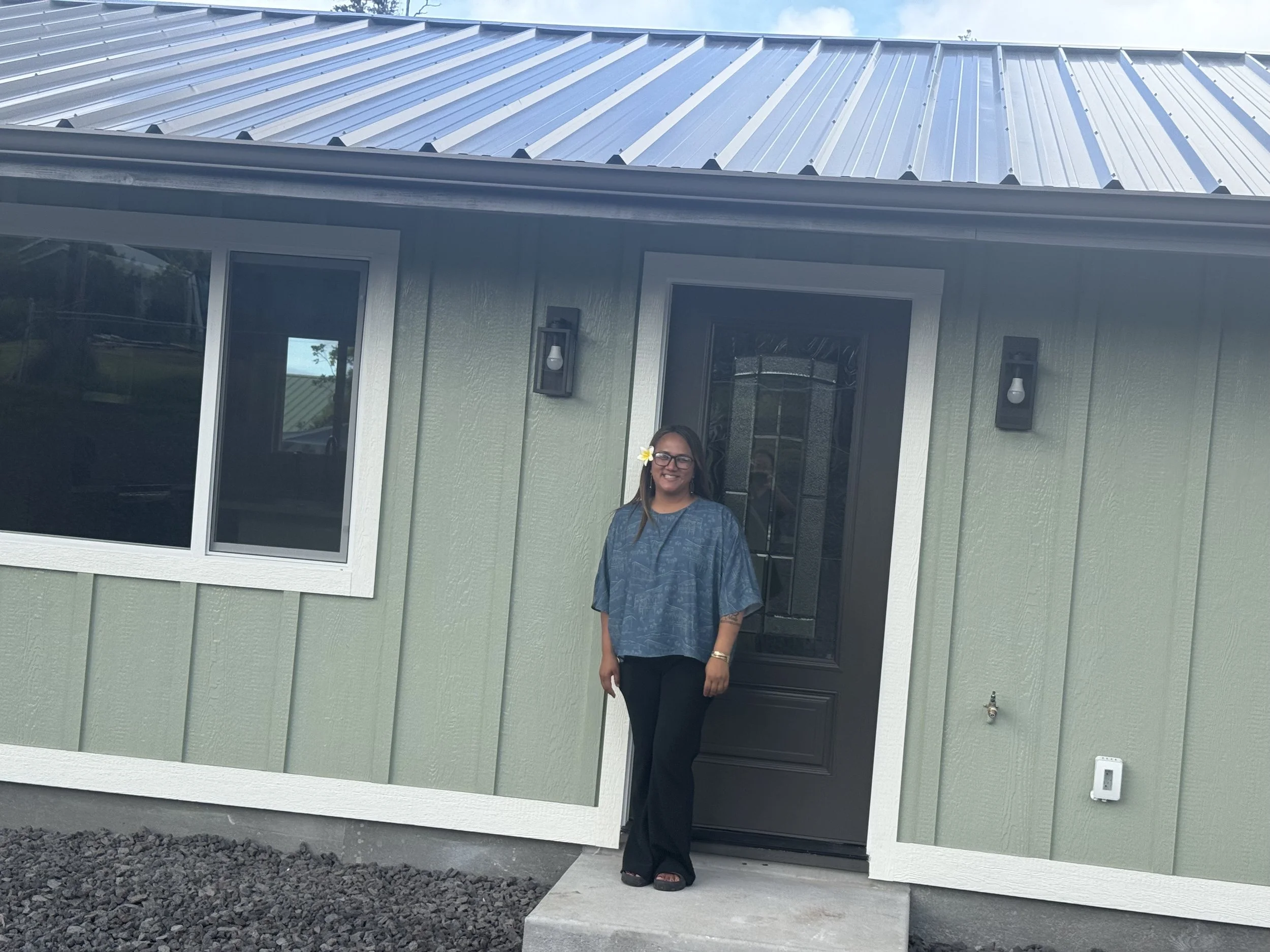 A woman with glasses standing in front of a green house with black door and windows, smiling, with a flower in her hair.