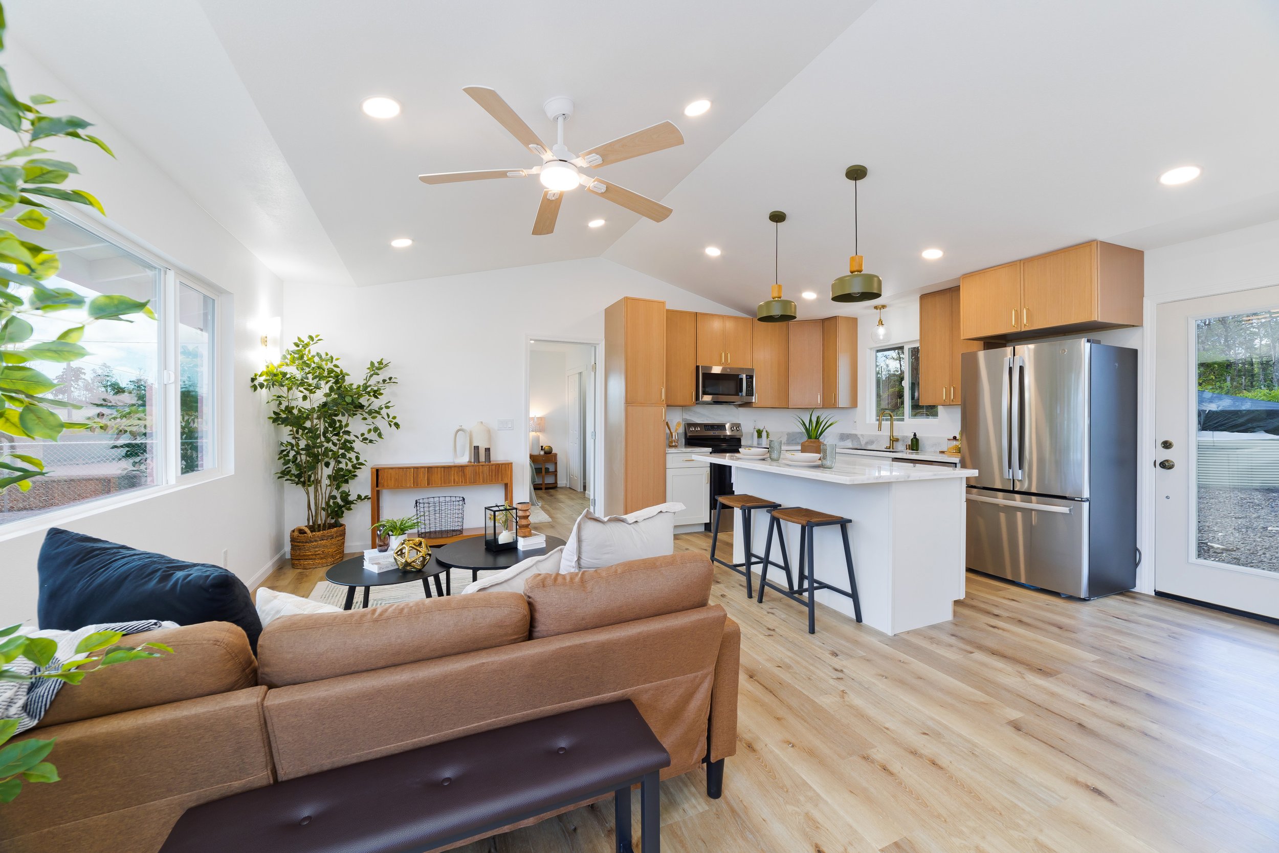 Open-concept living room and kitchen with hardwood floors, white walls, large window, ceiling fan, and modern appliances with wooden cabinets.