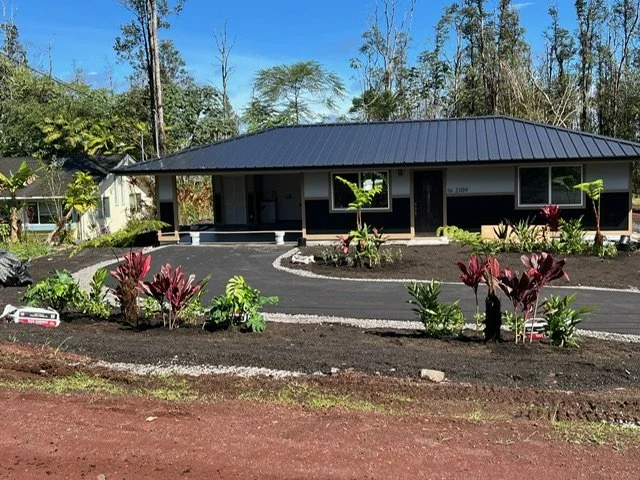 Single-story house with a black metal roof, surrounded by a landscaped yard with tropical plants and a paved driveway, in a rural area with trees and clear sky in the background.