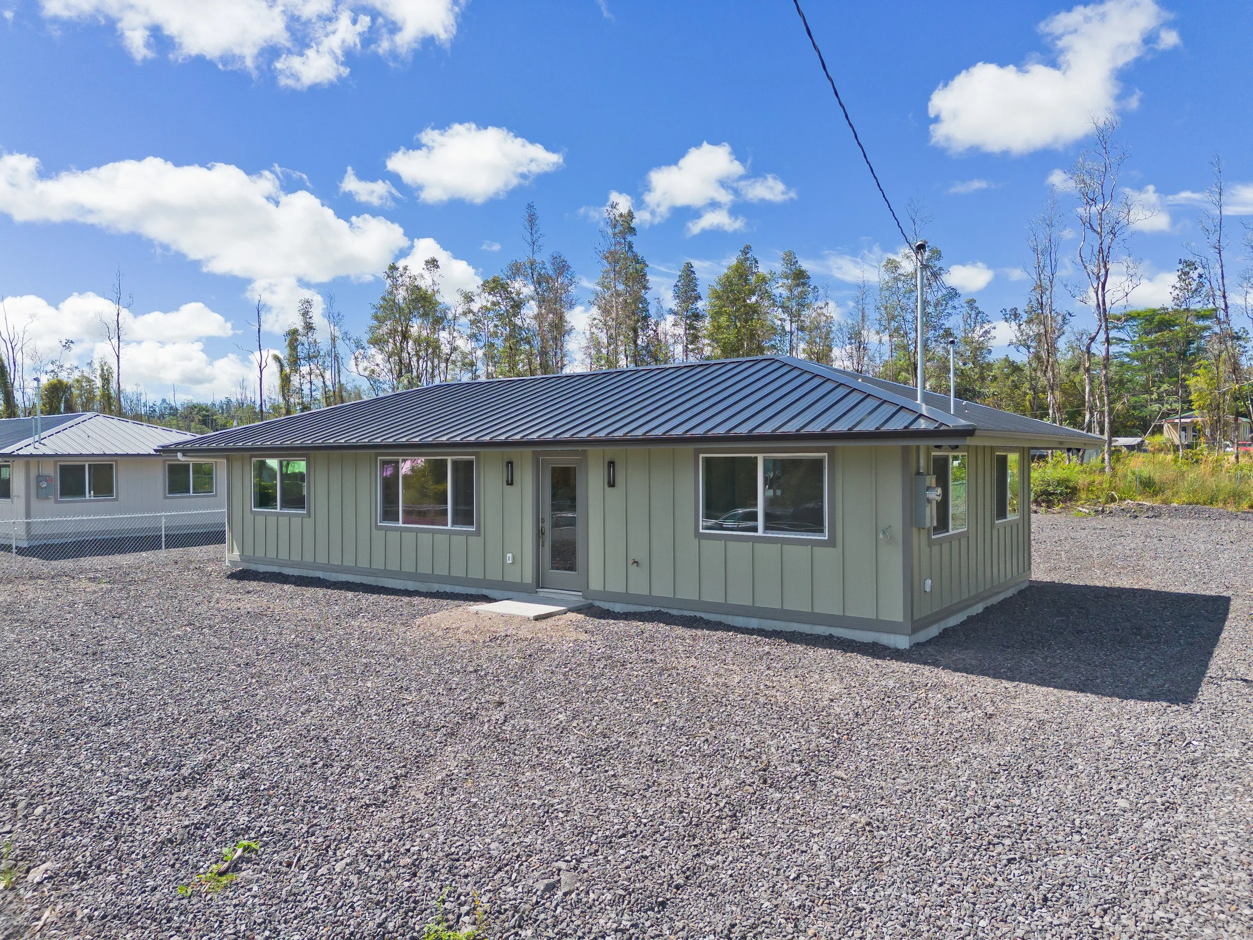 A newly built single-story house with beige siding, dark metal roof, and multiple windows, situated on a gravel lot under a partly cloudy sky.