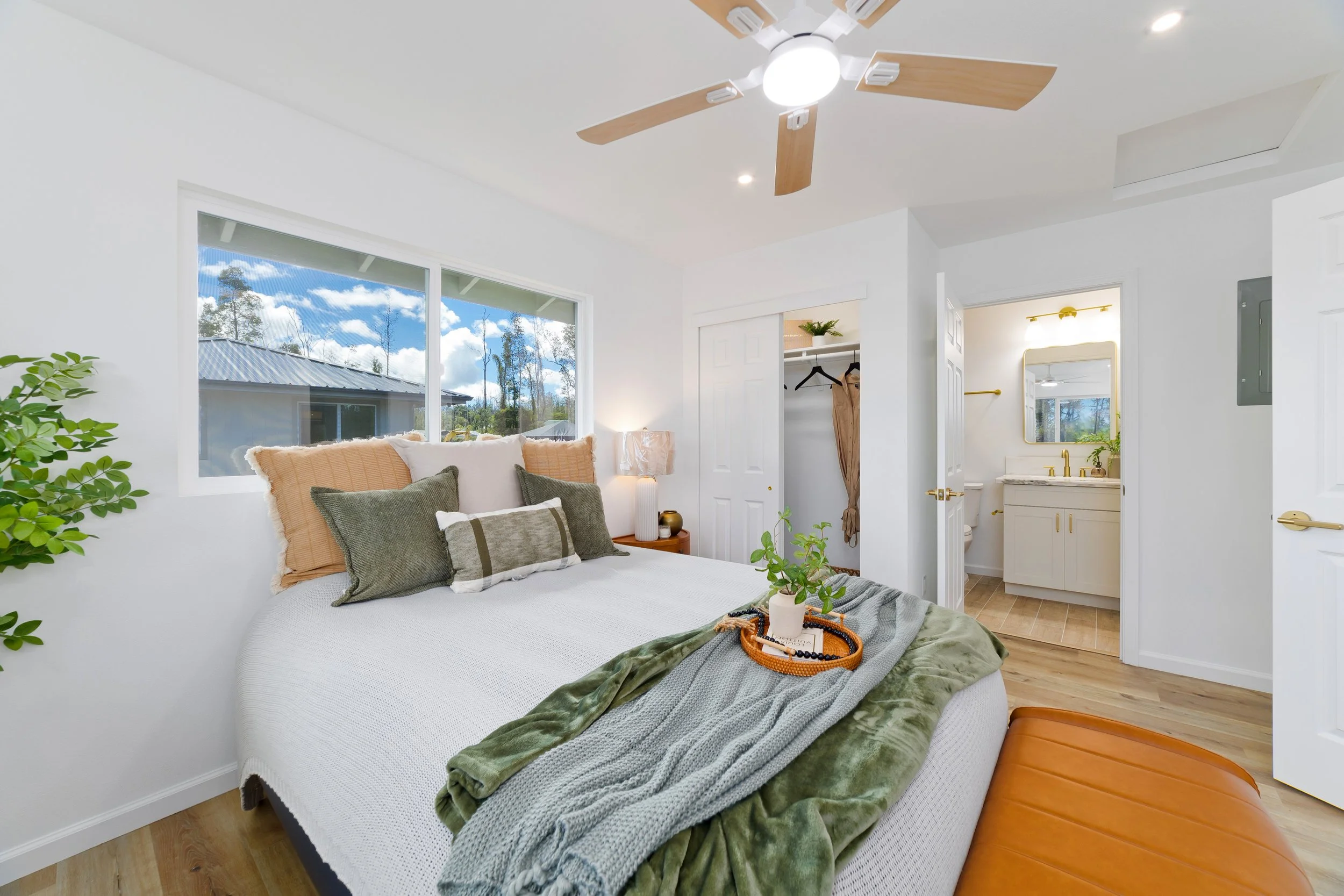Cozy bedroom with bed, pillows, blanket, plant, window showing outdoor trees, open closet, and attached bathroom with vanity.