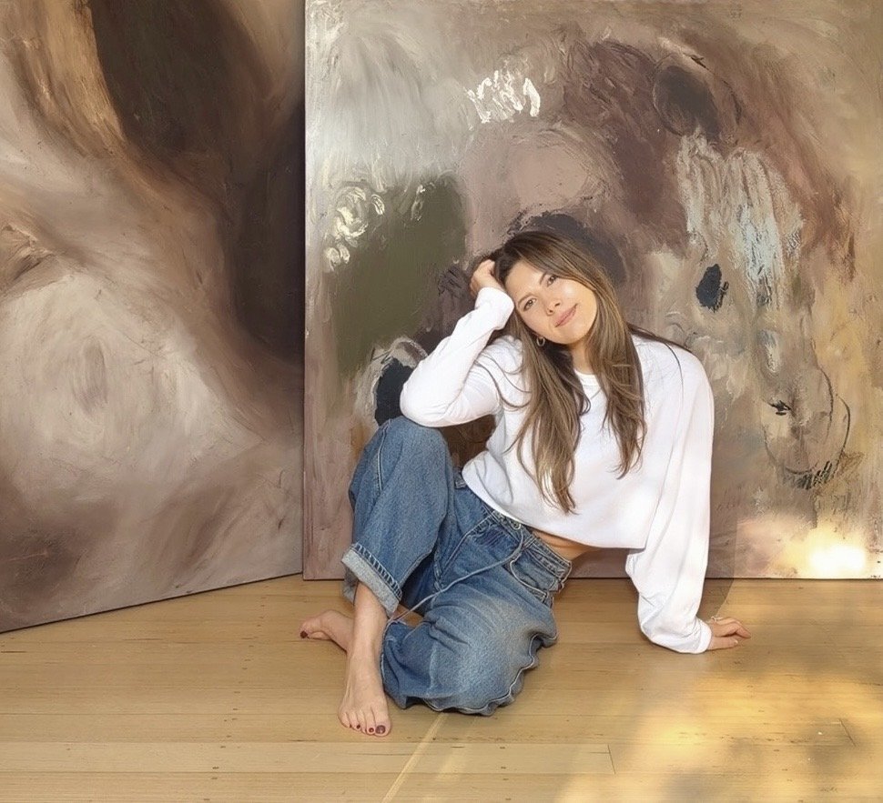 Young woman with long brown hair wearing a white long sleeve shirt and loose jeans, sitting on a wooden floor in front of large abstract paintings with brown and beige tones, posing with her hand on her head and smiling.