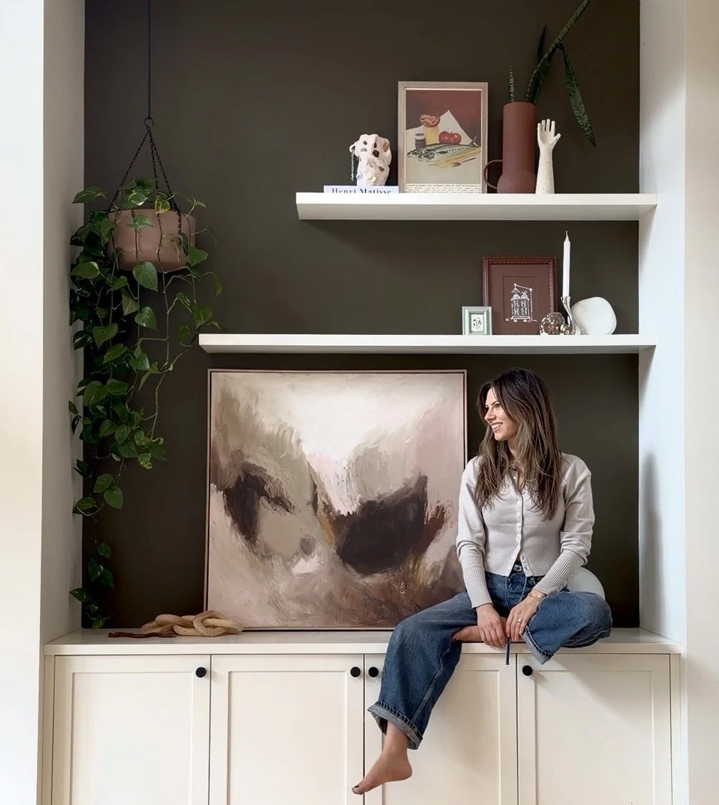 A woman with long brown hair, wearing a gray cardigan and blue jeans, sitting barefoot and smiling on a white cabinet in front of abstract artwork. The background features dark green wall with two white shelves decorated with plants, framed pictures, and decorative objects.
