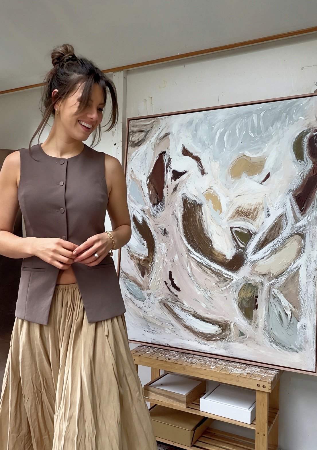 A woman standing in front of an abstract painting with swirling white, brown, and beige colors, smiling and looking down.
