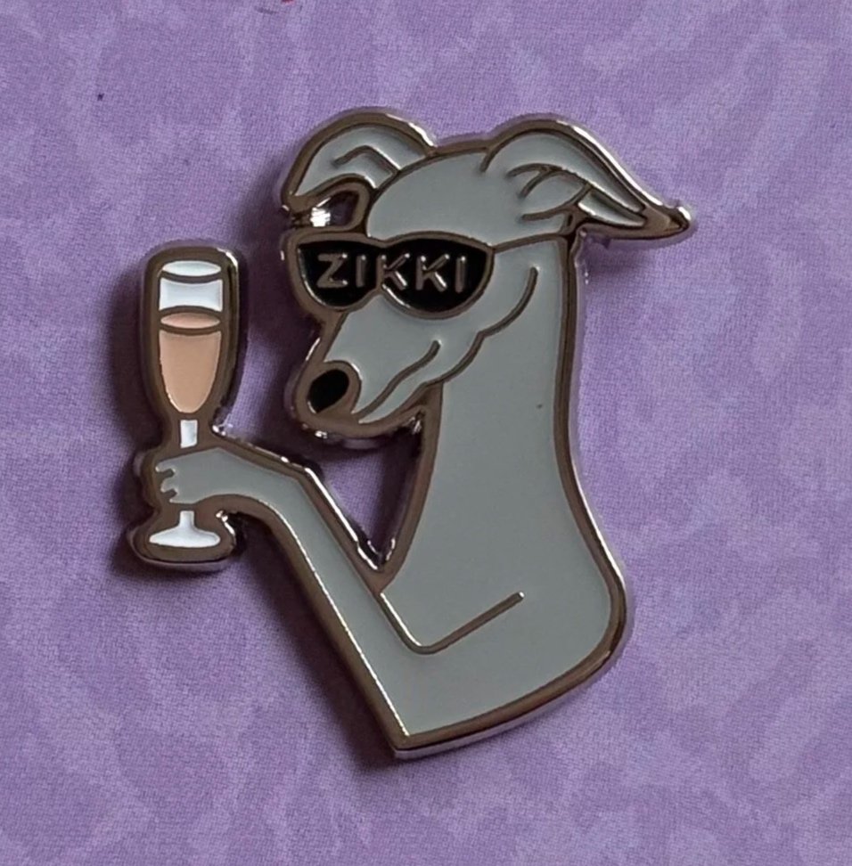 A silver-colored enamel pin of a dog wearing sunglasses labeled "ZIKKI", holding a champagne glass, placed on a purple fabric background.