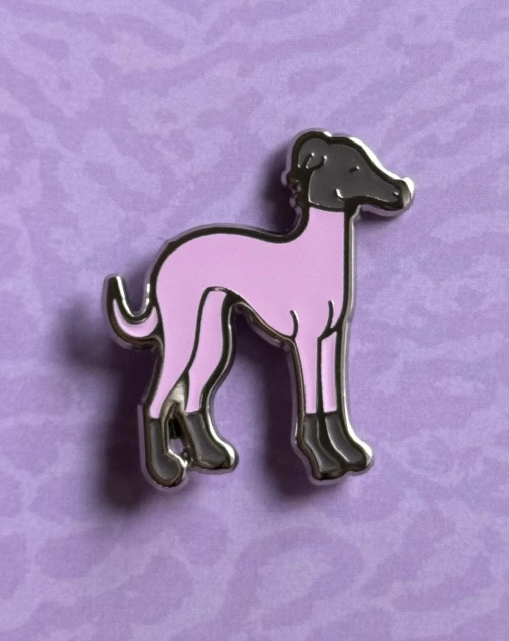 A pink and black enamel pin of a standing Italian Greyhound with a purple background.