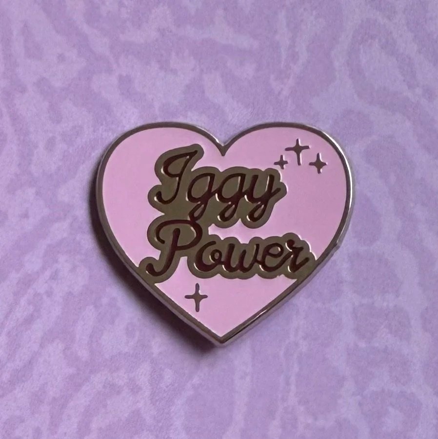 A purple heart-shaped pin with the words 'Gay Power' written in bold, black script, and small star-like decorations around the text.