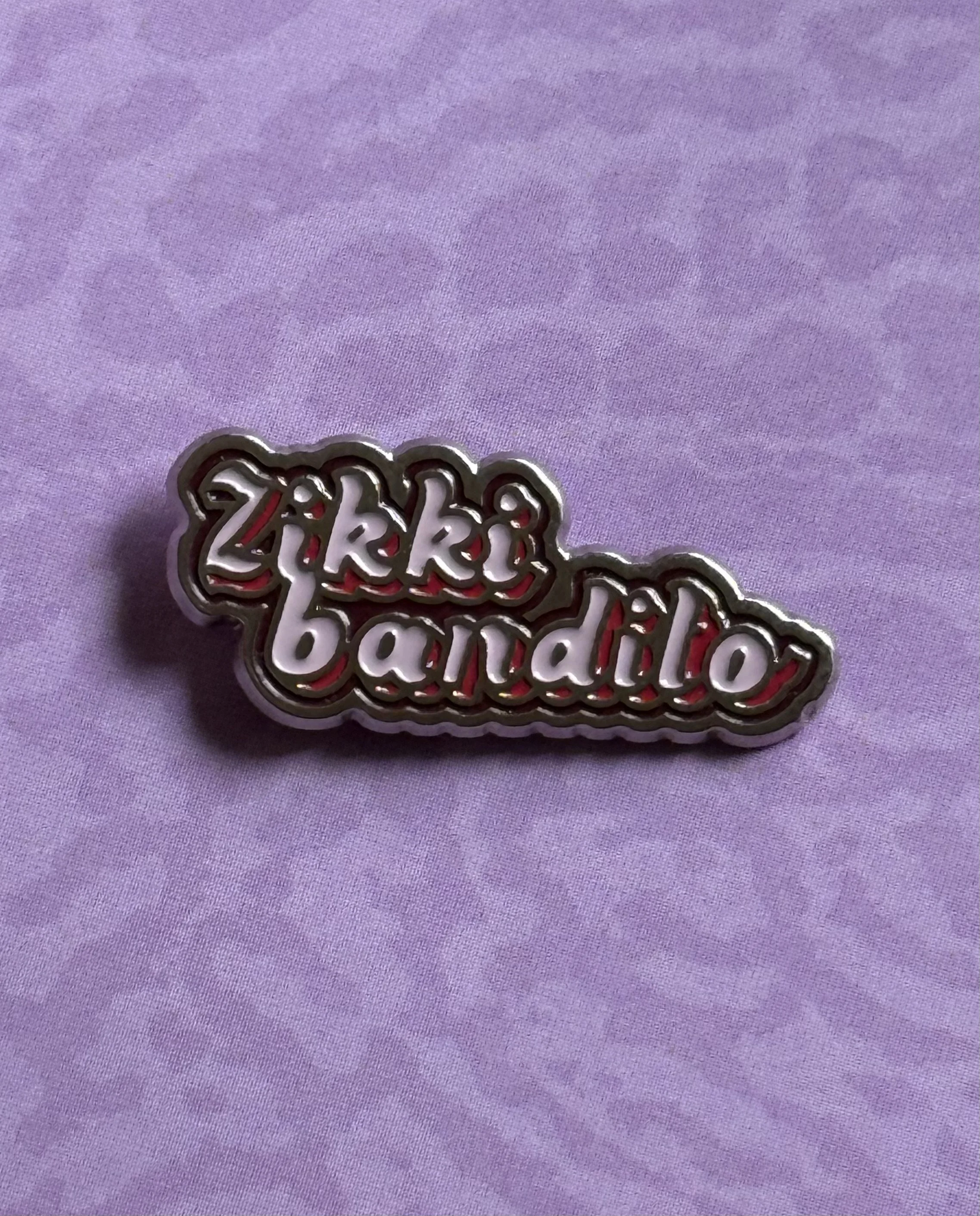 A colorful enamel pin with the words 'Zito Bandito' in a stylized font, placed on a purple fabric background.