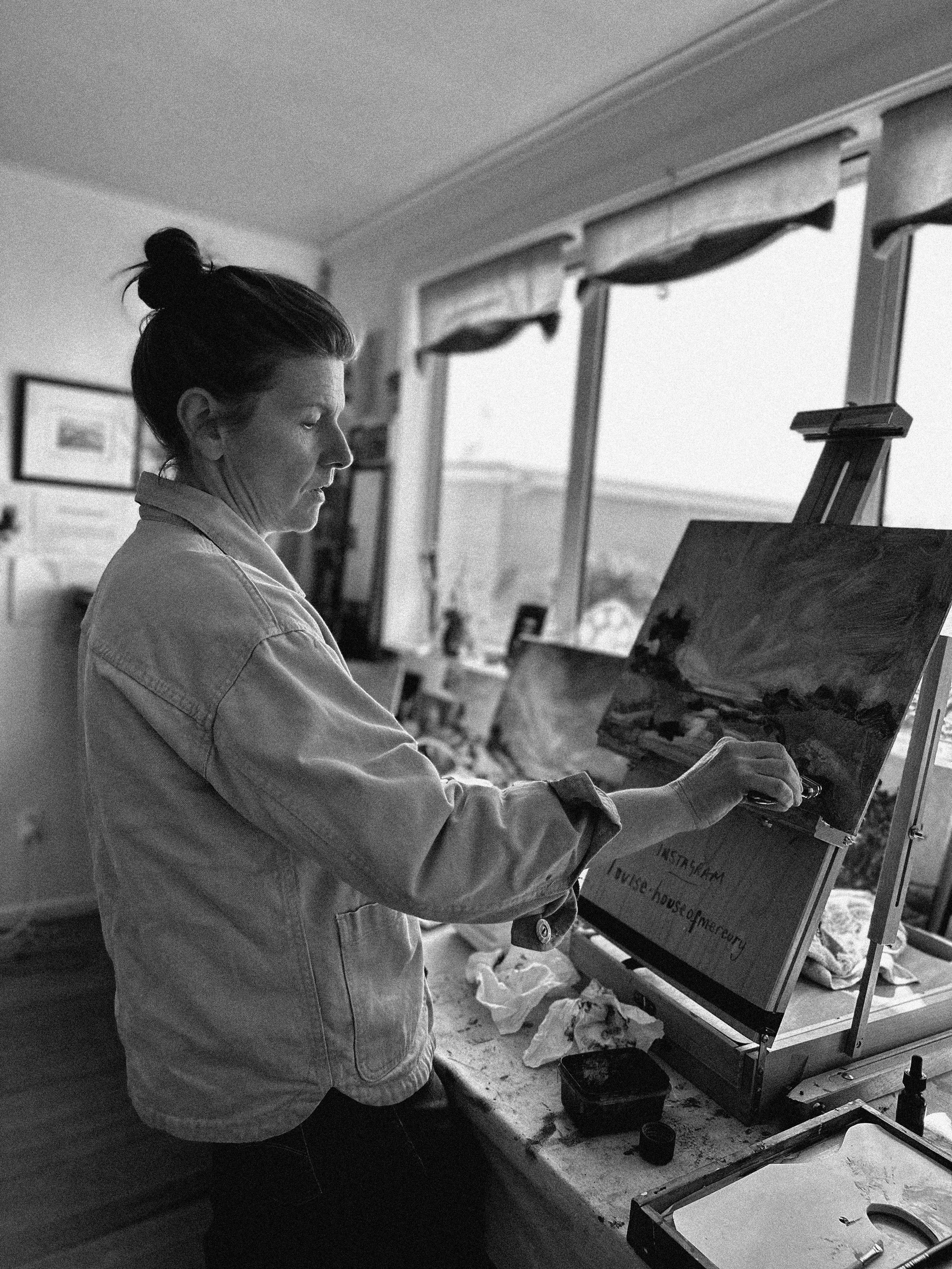 A person with dark hair in a bun painting on a canvas in a well-lit room with windows.