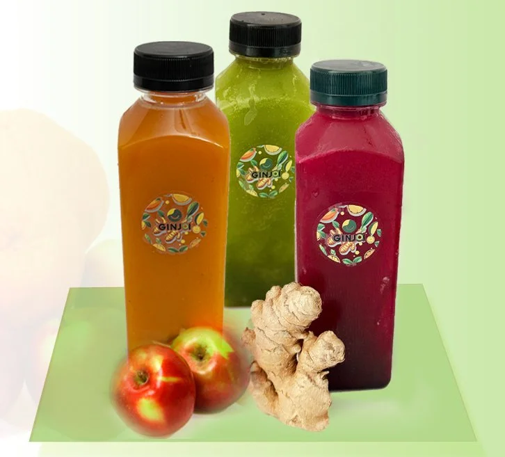 Three bottles of juice in orange, green, and pink colors with ginger and apples placed in front of them on a green surface with a light background.