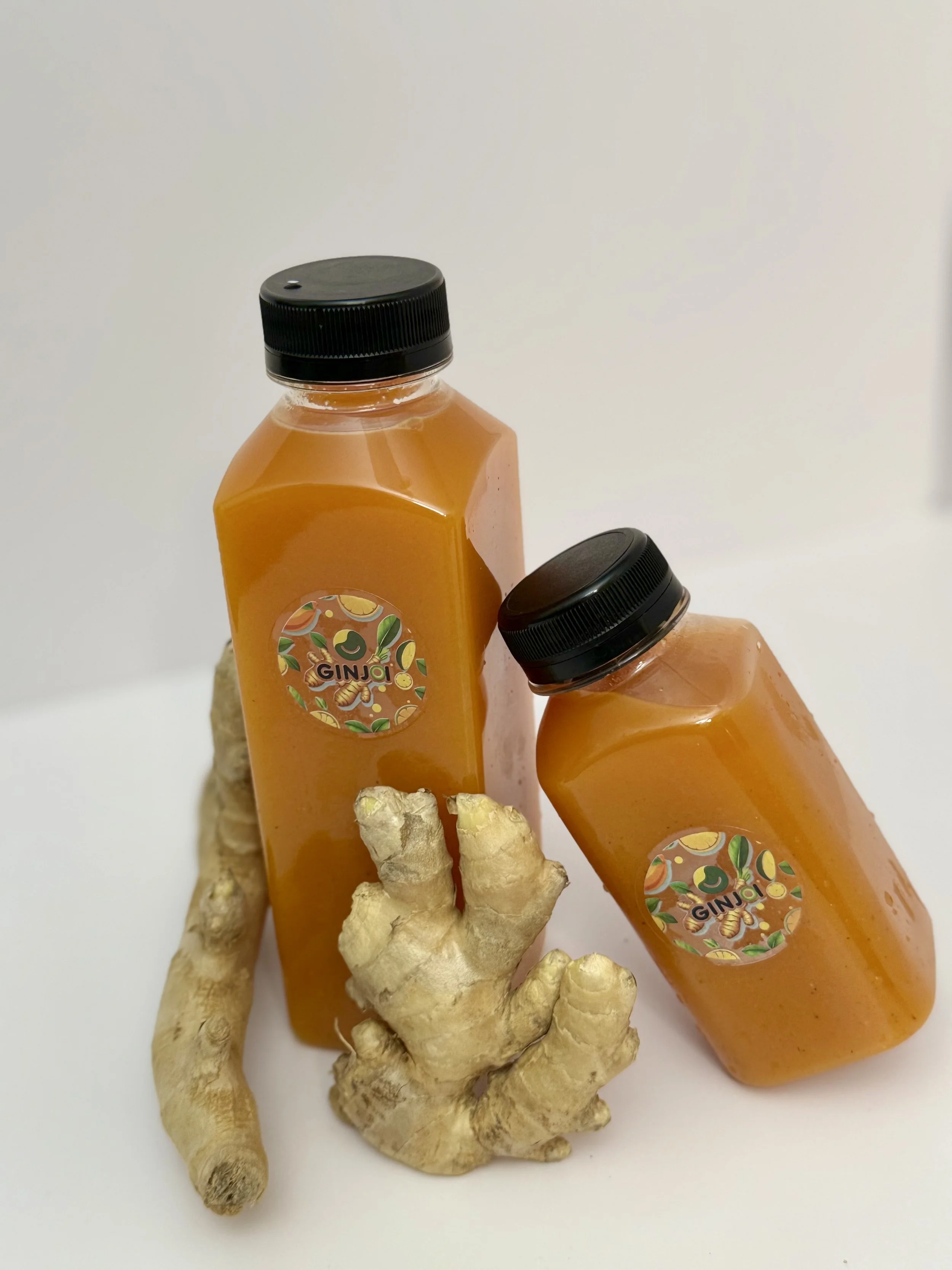 Two bottles of ginger juice with a ginger root and turmeric root in front, on a plain white background.