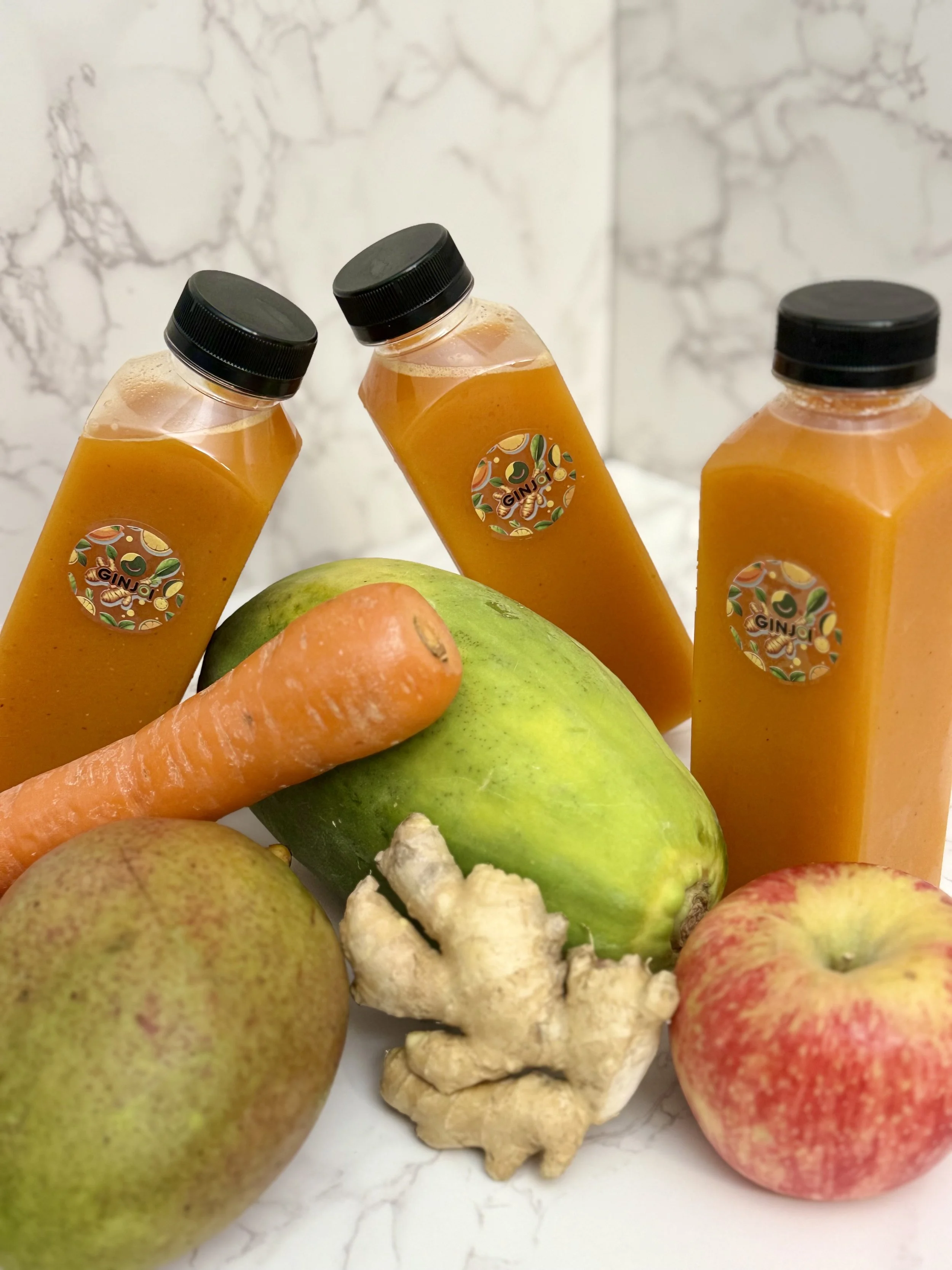 Three bottles of orange juice labeled 'GINJ', a carrot, a pear, a green apple, a piece of ginger root, and a red apple on a white marble surface.