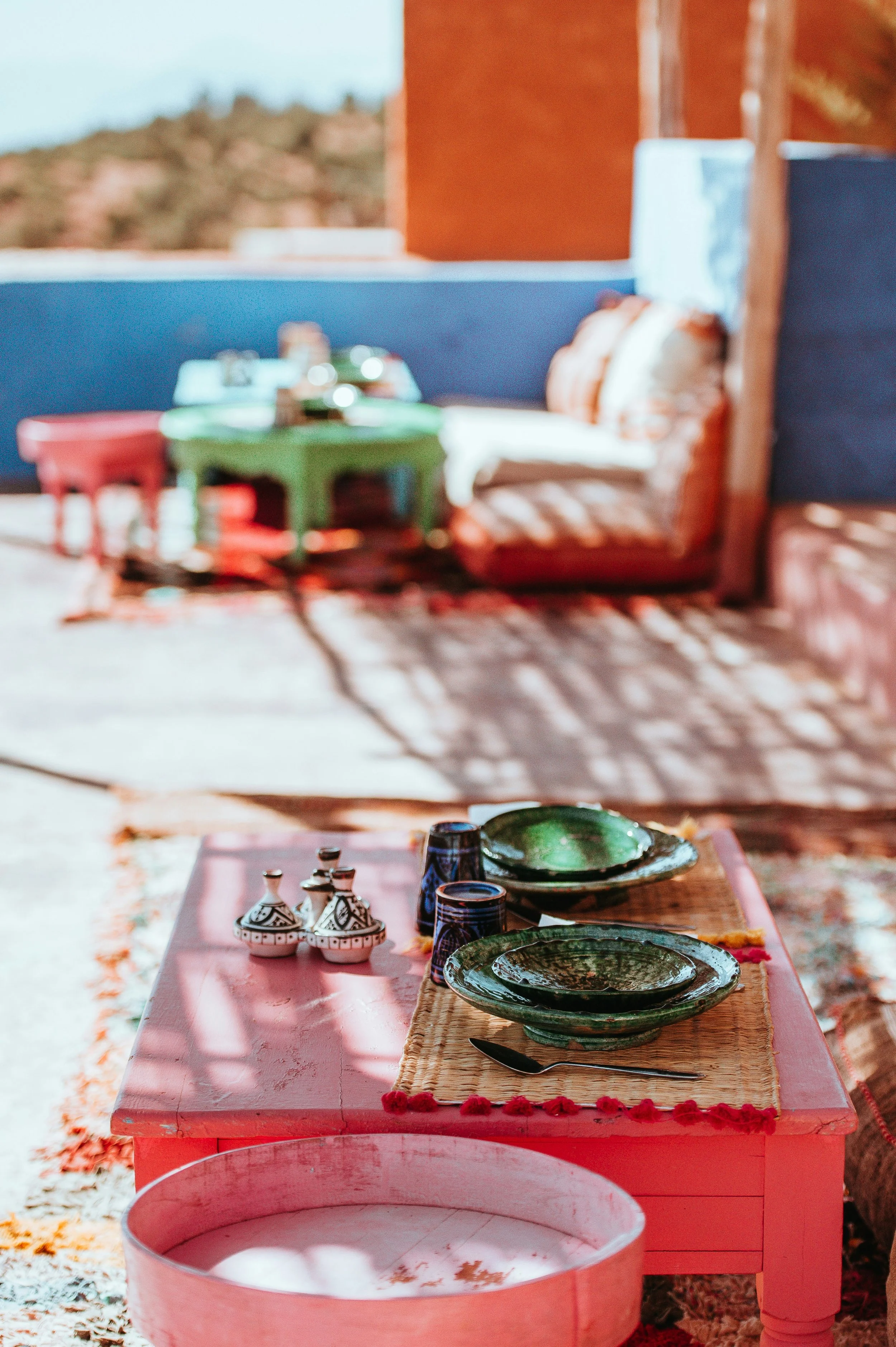 Outdoor patio table set for dining with green ceramic plates, blue cups, and a knife, with a pink table, and a second table with a green table and colorful chairs in the background.