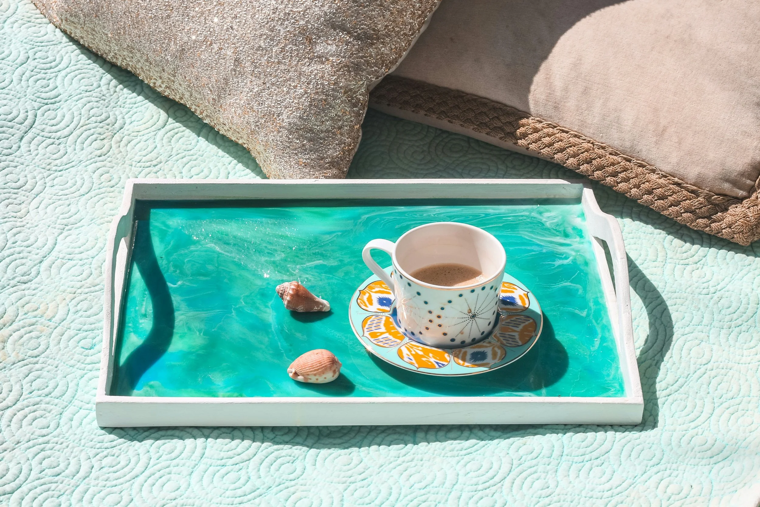 Tray with a cup of coffee, seashells, and a small seashell on a turquoise water surface, with pillows and a quilted blanket in the background.