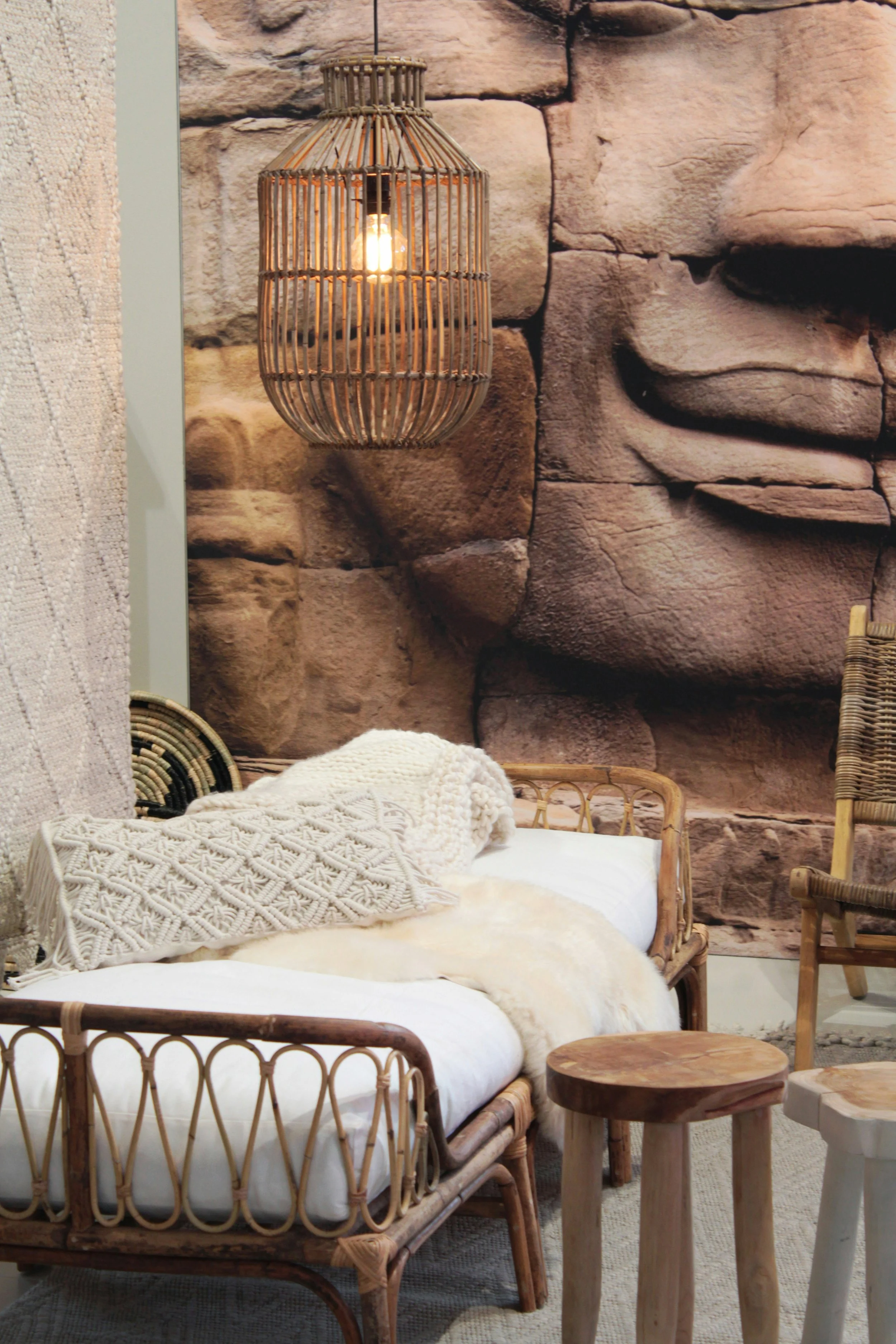 Wooden wicker pendant light hanging above a cozy rattan bed with white bedding, decorative pillows, and a fur throw, against a rocky stone wall with a large carved face detail.